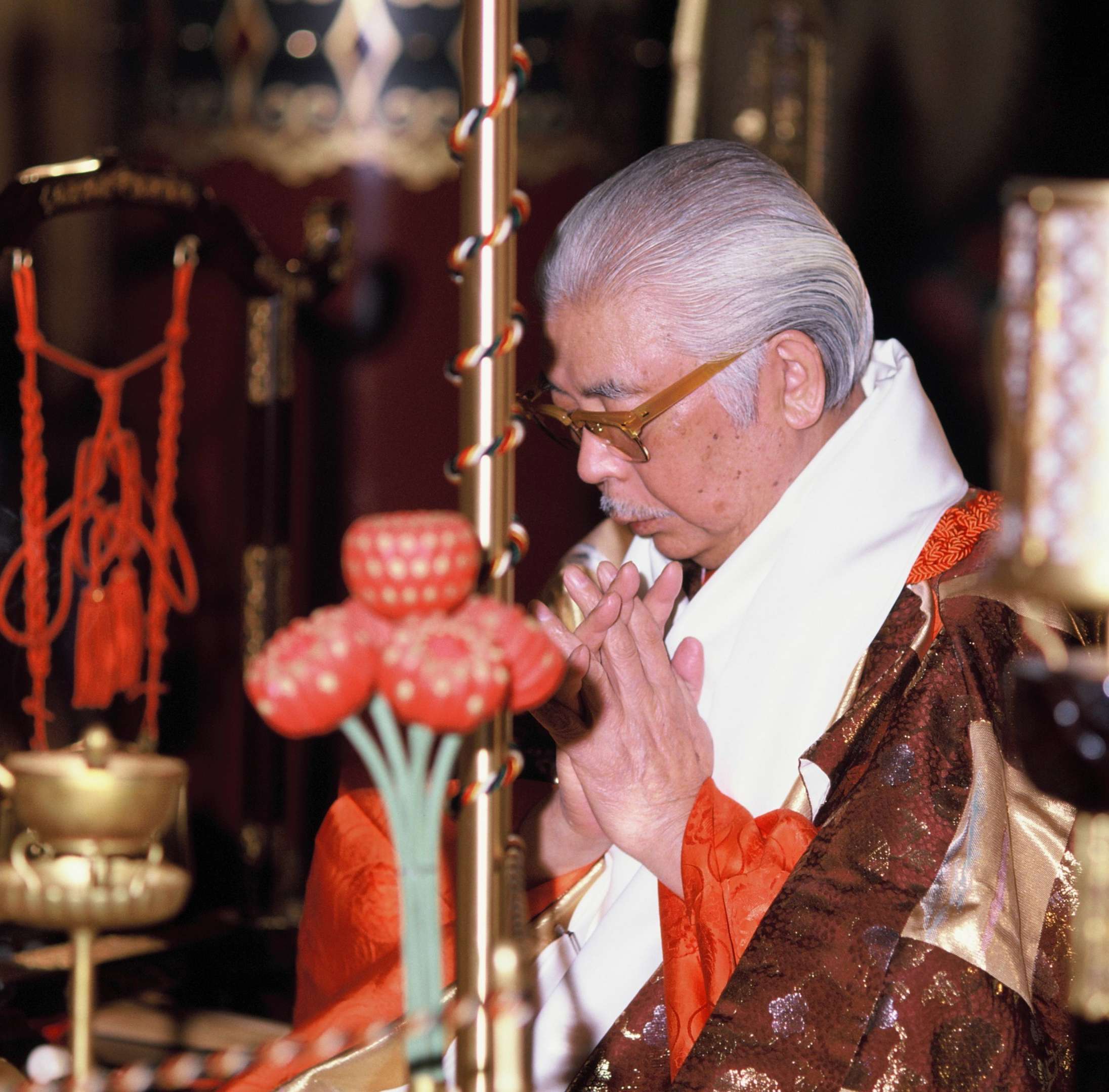 An elder Japanese man with white and gray hair and a trimmed moustache, wearing brown and vermillion brocade vestments, folds his hands and bows his head in prayer before a ritual altar on which can be seen golden implements and intricately crafted wooden lotuses.