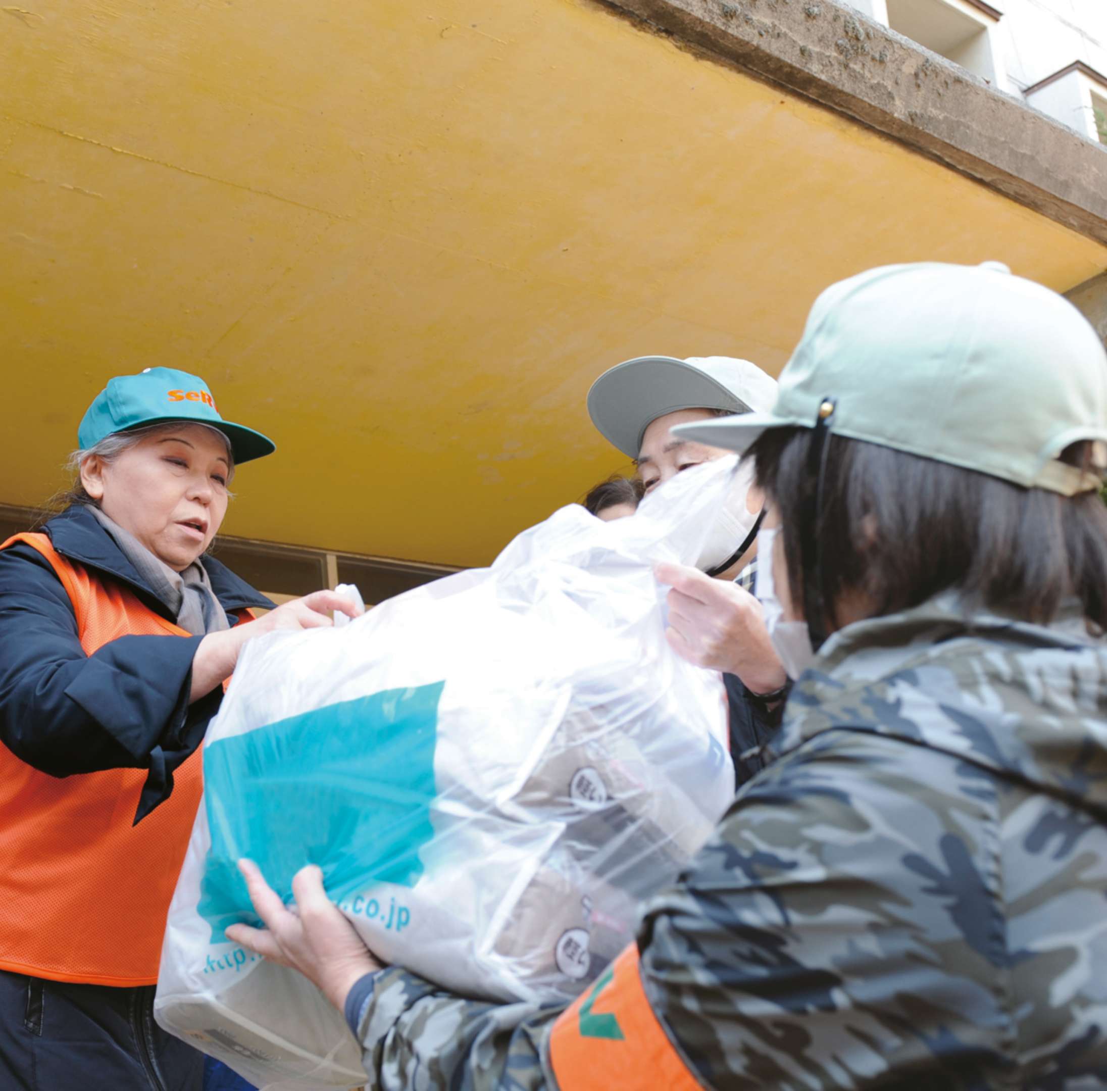 Her Holiness, wearing an orange safety vest and SEVA ballcap, passes a bag of relief supplies to people wearing caps and masks.