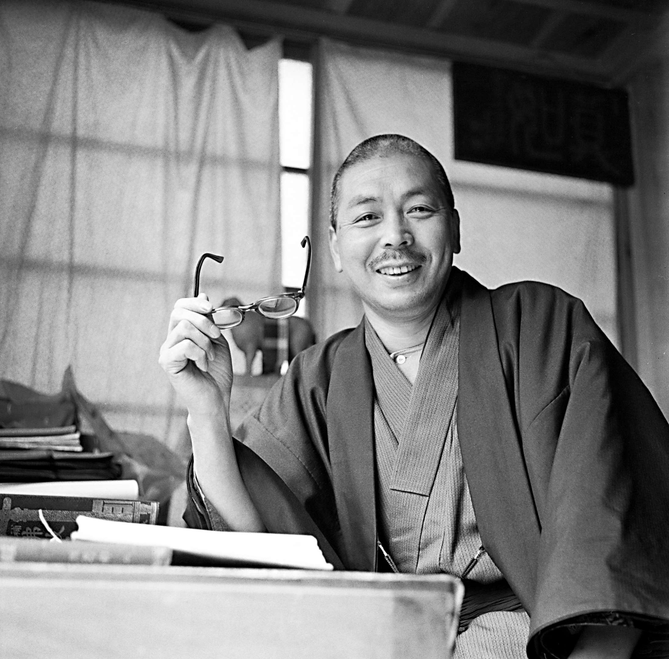 A candid photograph of Shinjo smiling in his study, shaven headed, mustachioed, in casual kimono, with elbow resting on table and reading glasses in hand.
