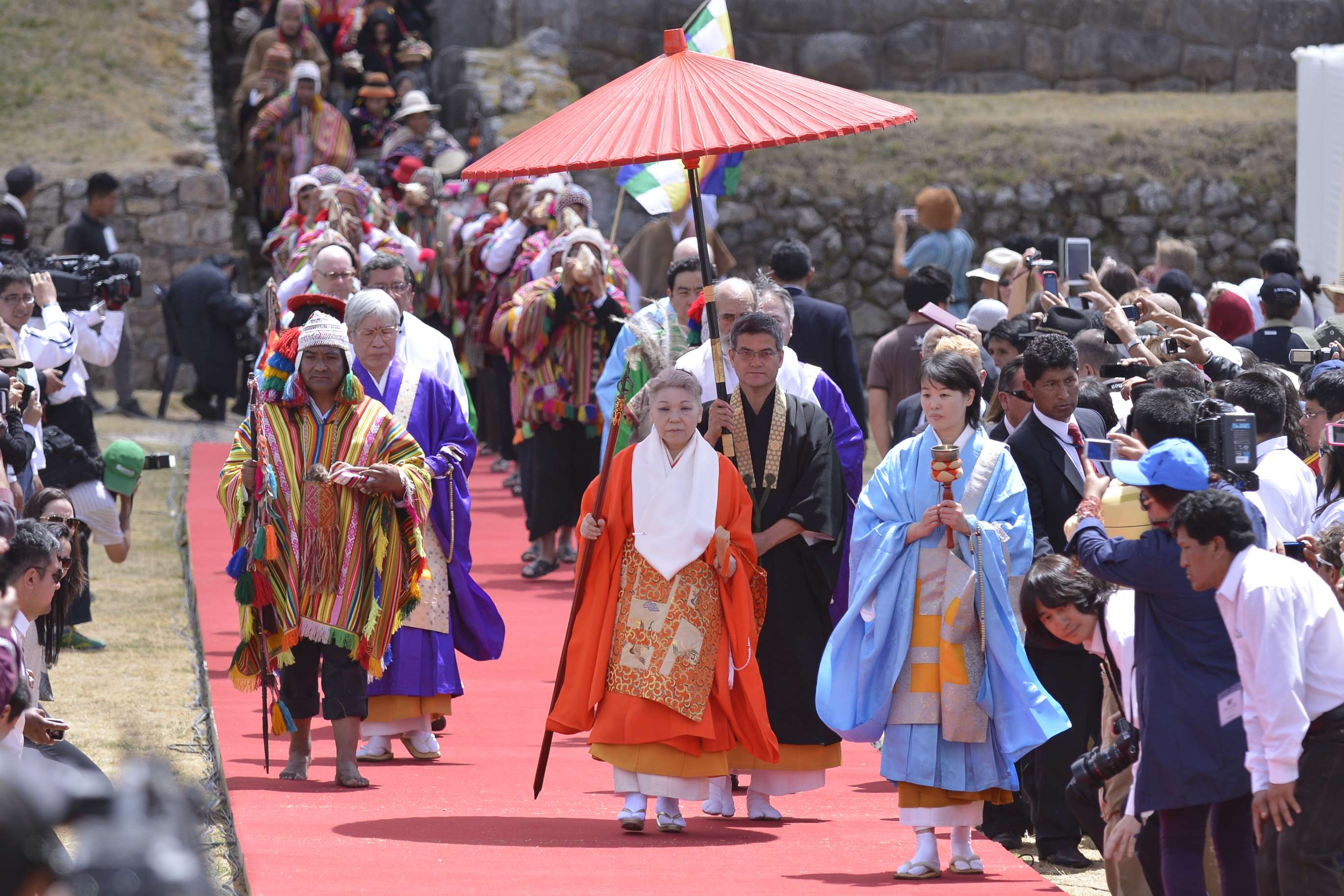 Her Holiness in ceremonial robes, carrying a staff, walks down an red-colored outdoor walkway accompanied by priests in robes and a Peruvian man in colorful native dress as a crowd looks on.