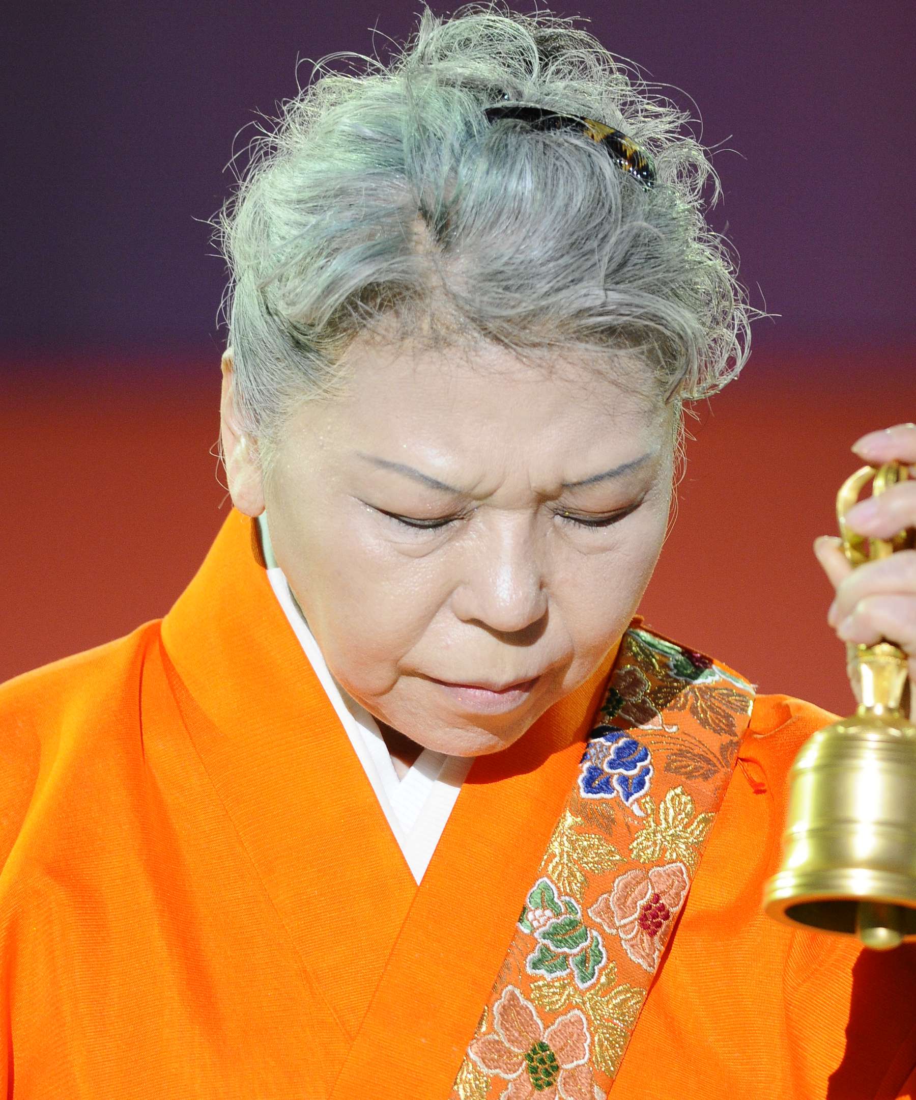 A photo of Her Holiness, gray-haired, wearing a bright orange robe with floral hem, eyes closed in deep concentration, holding a golden hand bell while performing a ritual.