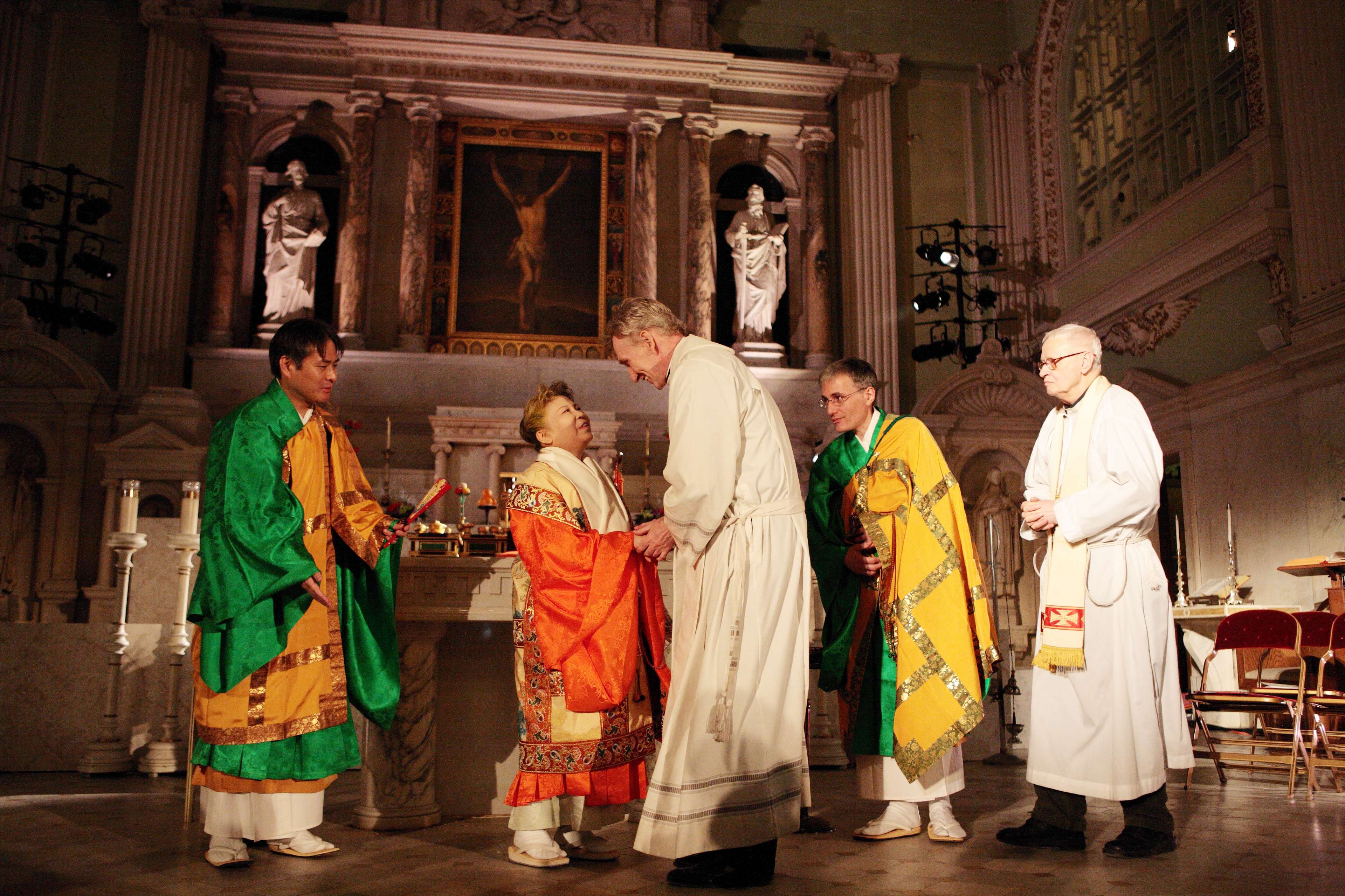 In a large cloister with Catholic statues, Her Holiness, in orange robe, embraces the hands of a tall Catholic priest as they both look smilingly at each other; three other priests stand watching.