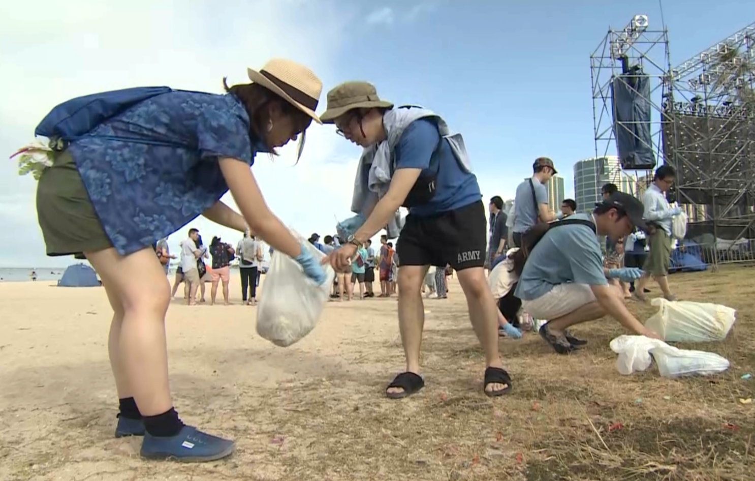 Two people wearing plastic gloves and carrying plastic bags stoop to pick up trash from the sandy ground; similarly active people are visible in the background.