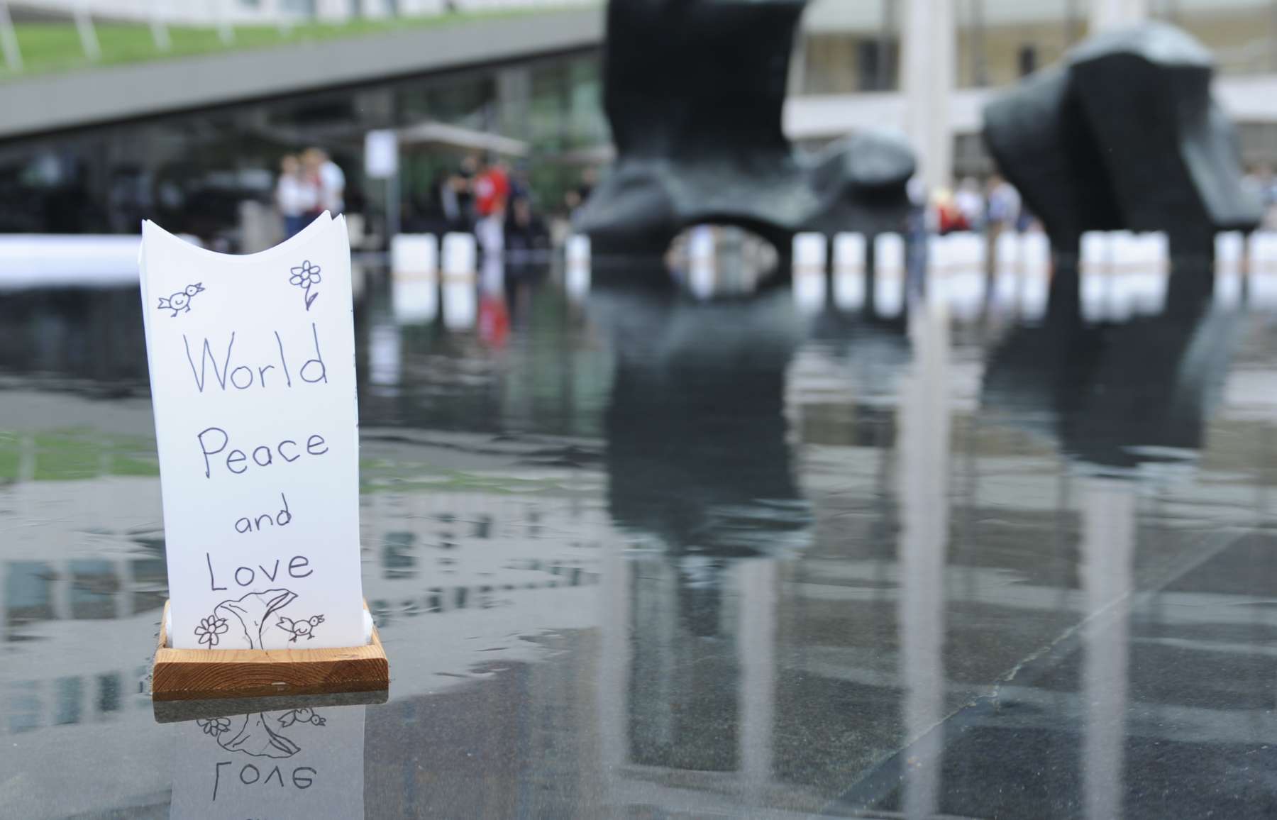 A simple lantern of paper with a wooden base with a written message reading “World Peace and Love” can be seen, floats in a still body of water, causing ripples.