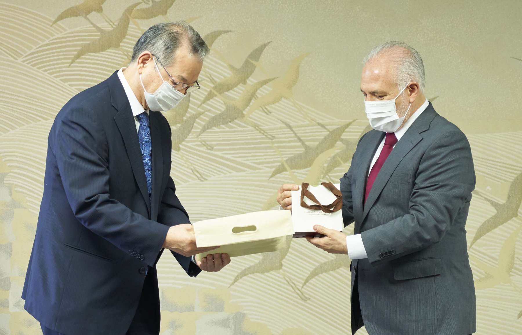 Two men wearing suits and face masks pass a formal document one to the other in front of a background illustration of cranes taking flight.