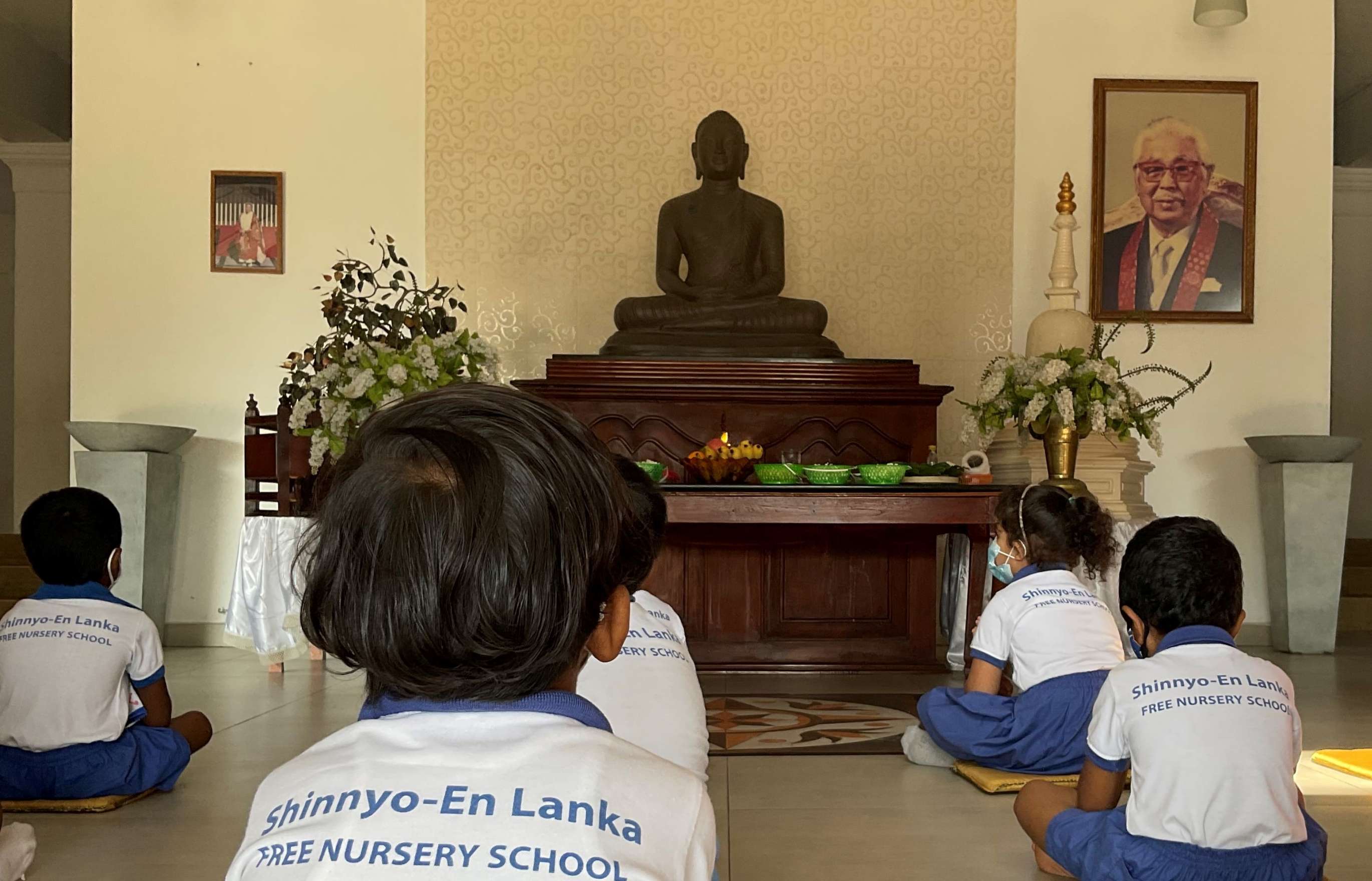 A group of children wearing blue shorts and tee-shirts with the words “Shinnyo-en Lanka Nursery School” printed on them sit mindfully in front of a statue of Buddha.