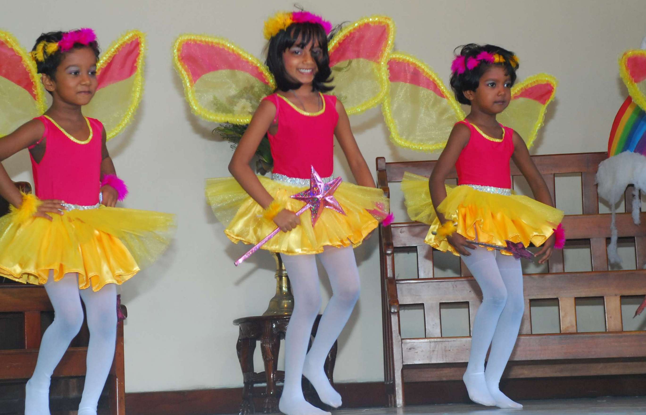 Three girls in yellow and pink tutus, white tights, and wearing yellow and pink butterfly wings perform a dance.
