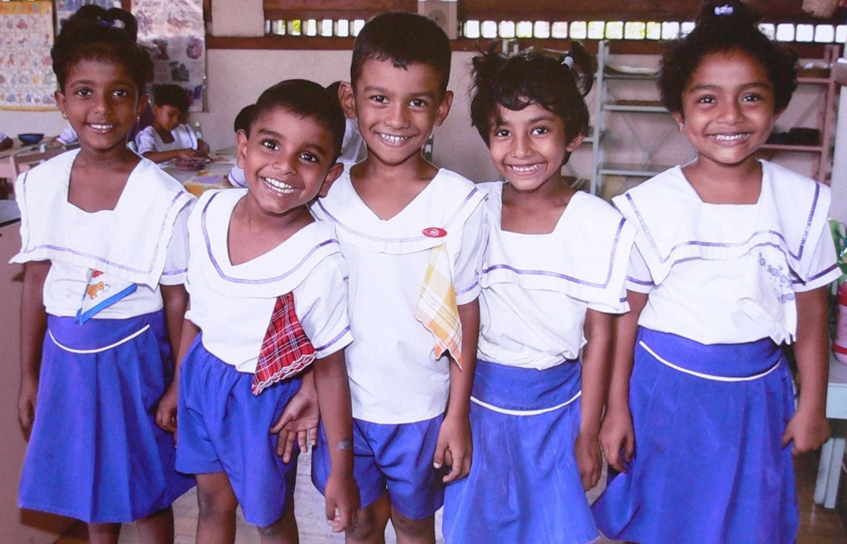 Five smiling school children in blue and white school uniforms pose side-by-side for a photograph.