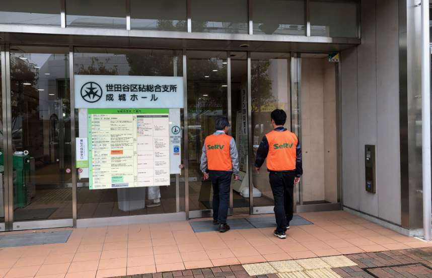 Two men wearing orange SeRV volunteer vests over suits approach the glass doors at the entry to a building with a large sign in Japanese next to the door.