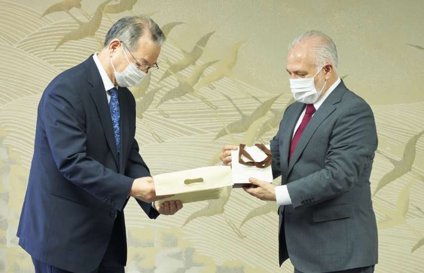 Two men wearing suits and face masks pass a formal document one to the other in front of a background illustration of cranes taking flight.