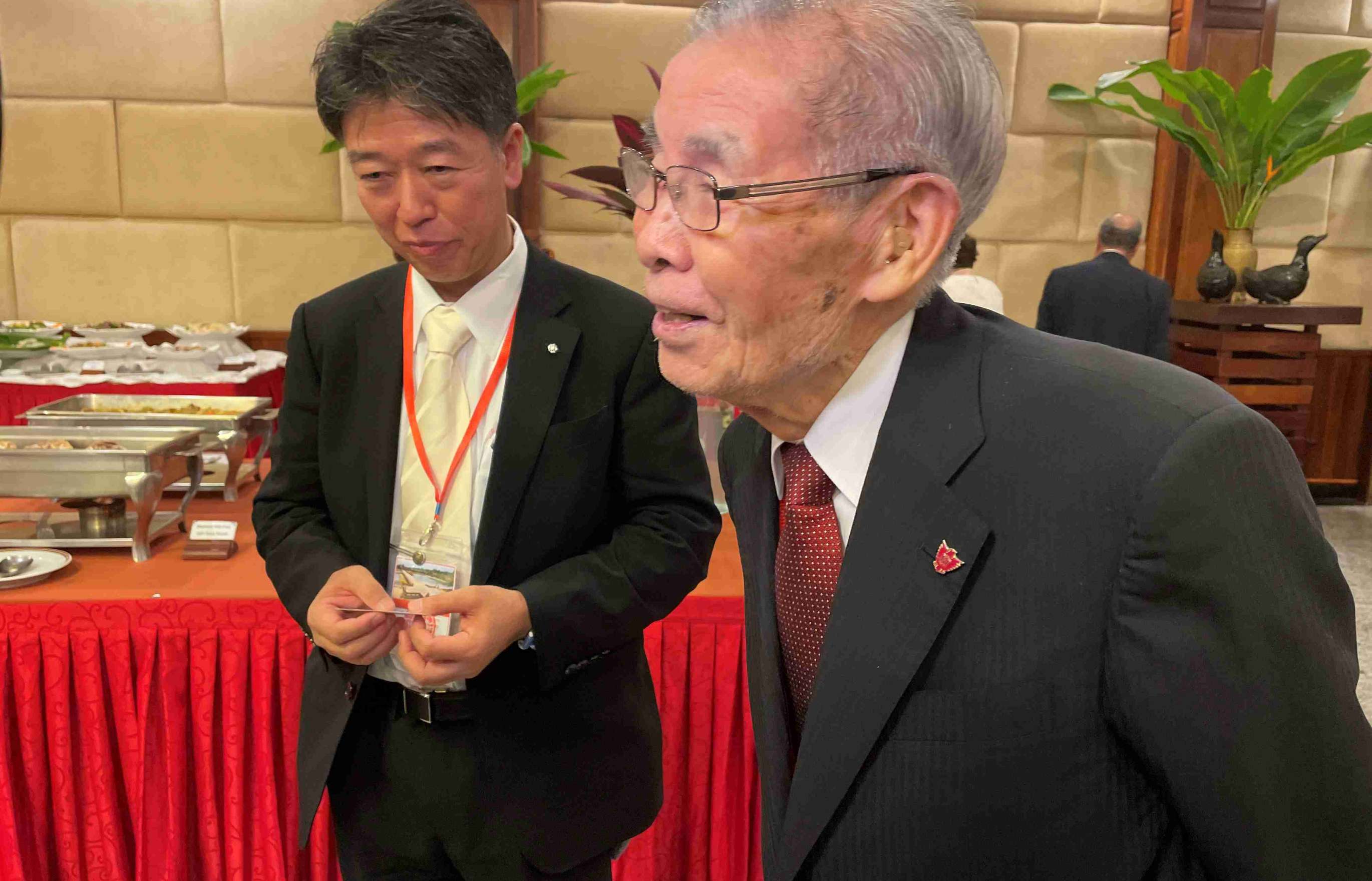 An elderly, distinguished looking, bespectacled Japanese man in a charcoal gray suit with his arms folded behind his back speaks to someone out of frame in a banquet hall. A younger Japanese man in a suit in the background listens to the conversation.