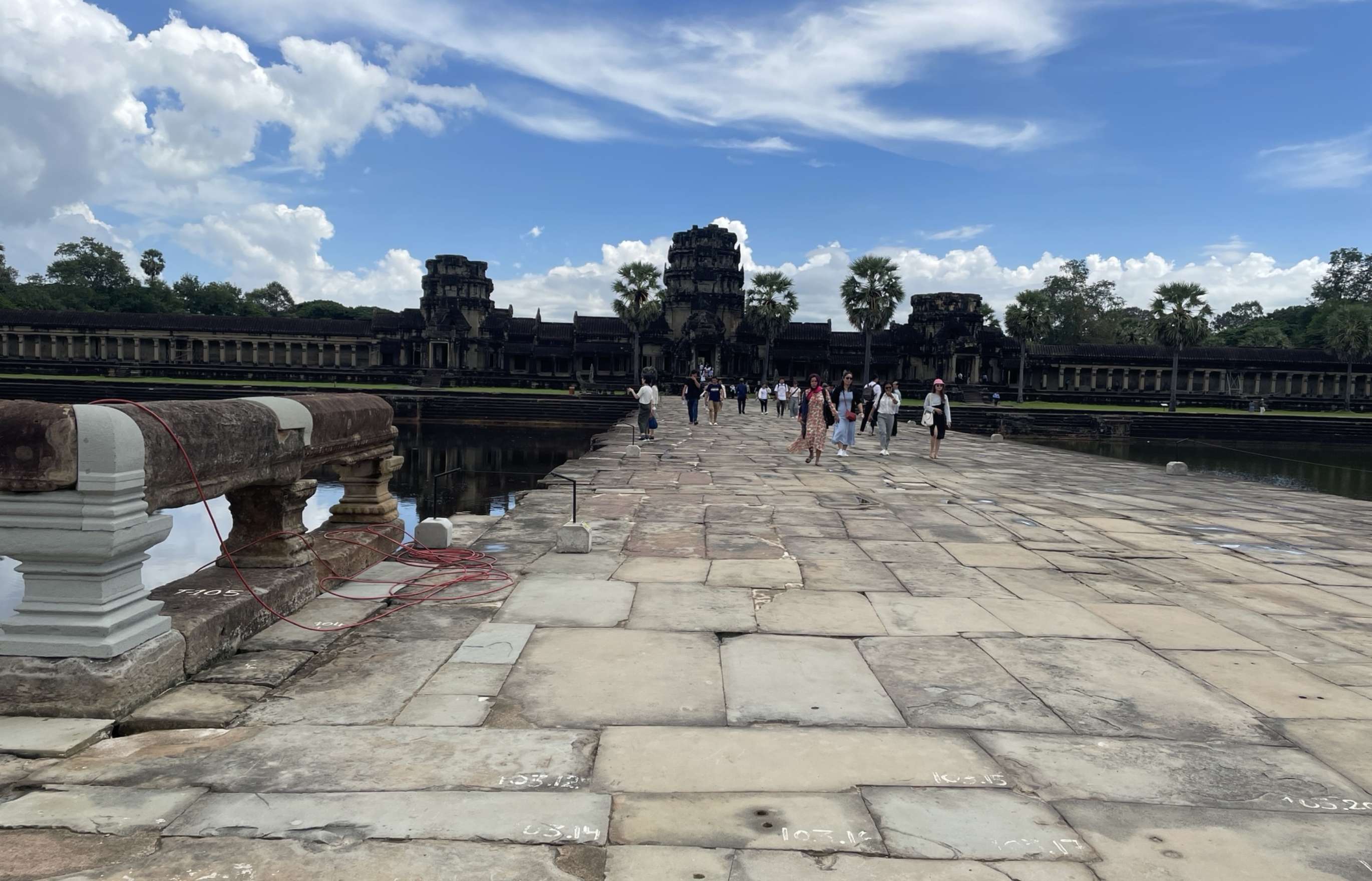 The camera looks down a long causeway across a large moat toward a temple ruin rising from the palms beneath a blue, cloud dappled sky in the distance. Small groups of people are visible far down the causeway, which is paved with giant slabs of stone.