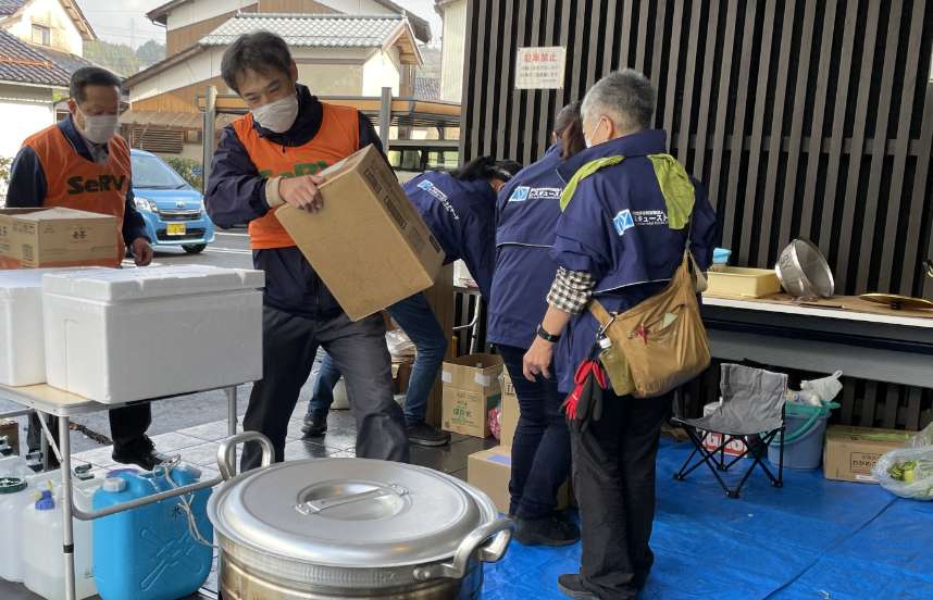 At the entrance to a building, a man wearing an orange hi-vis vest with a SeRV logo printed on the back of it carries a large white box toward a woman waiting to receive it with outstretched arms; large empty water containers, trash bags, and a makeshift station for distribution of food and water are visible in the scene.