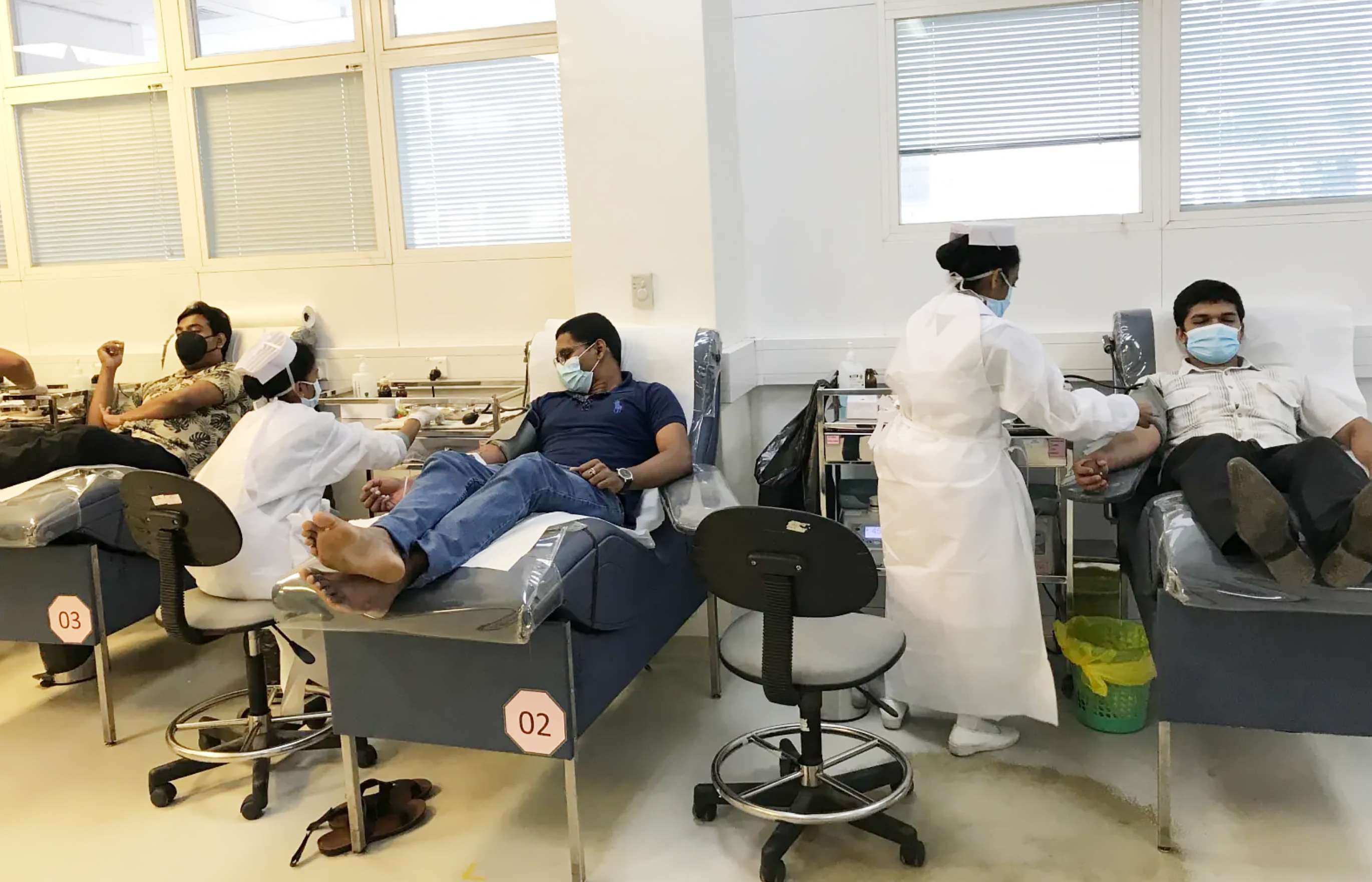 Three men wearing masks are in a reclined position on a chair having their blood drawn by nurses wearing white gowns.