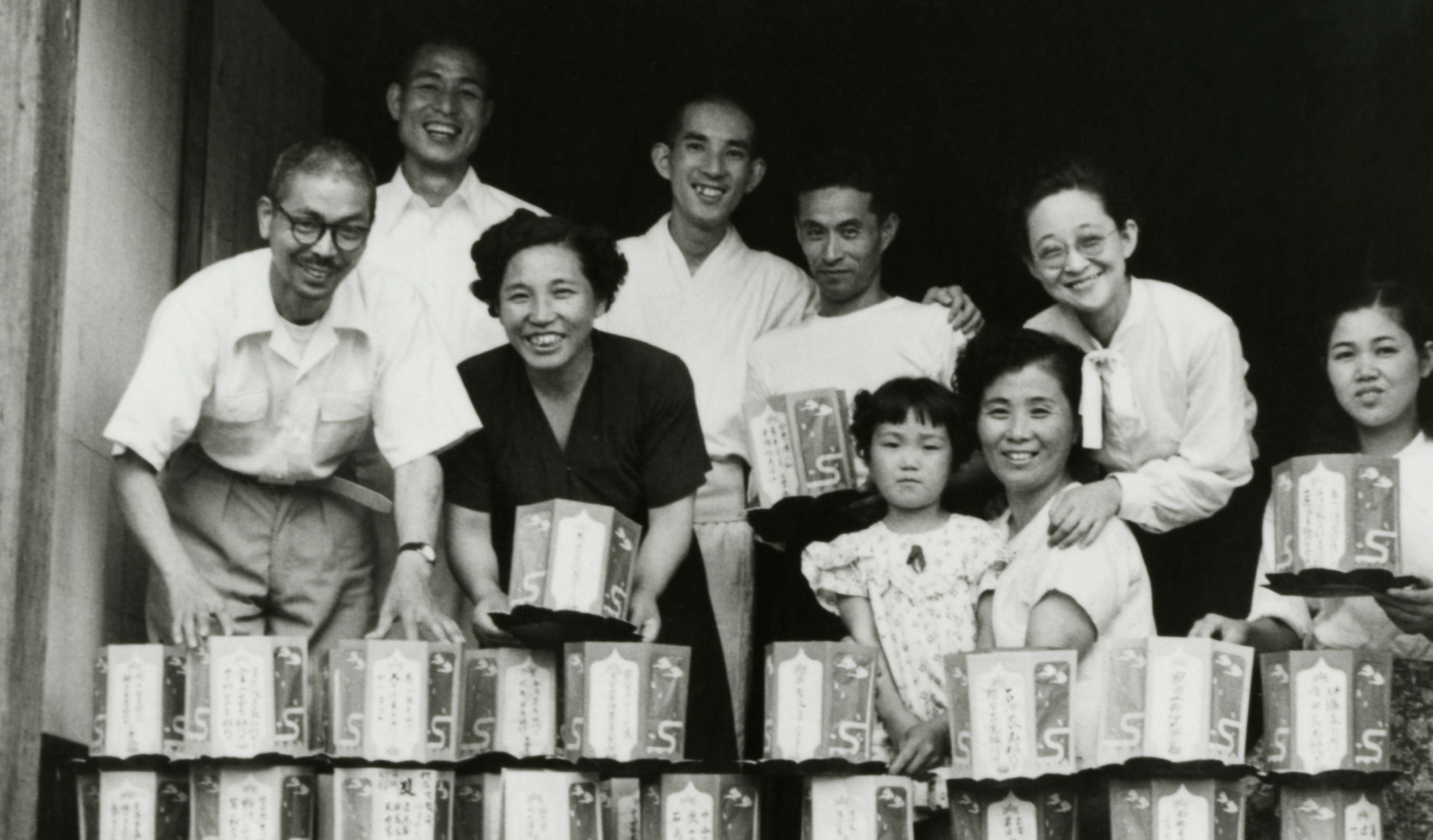 A beaming Shinjo and Tomoji, wearing lay clothes, pose for a portrait with a smiling group of men, women, and children preparing and stacking paper lanterns.