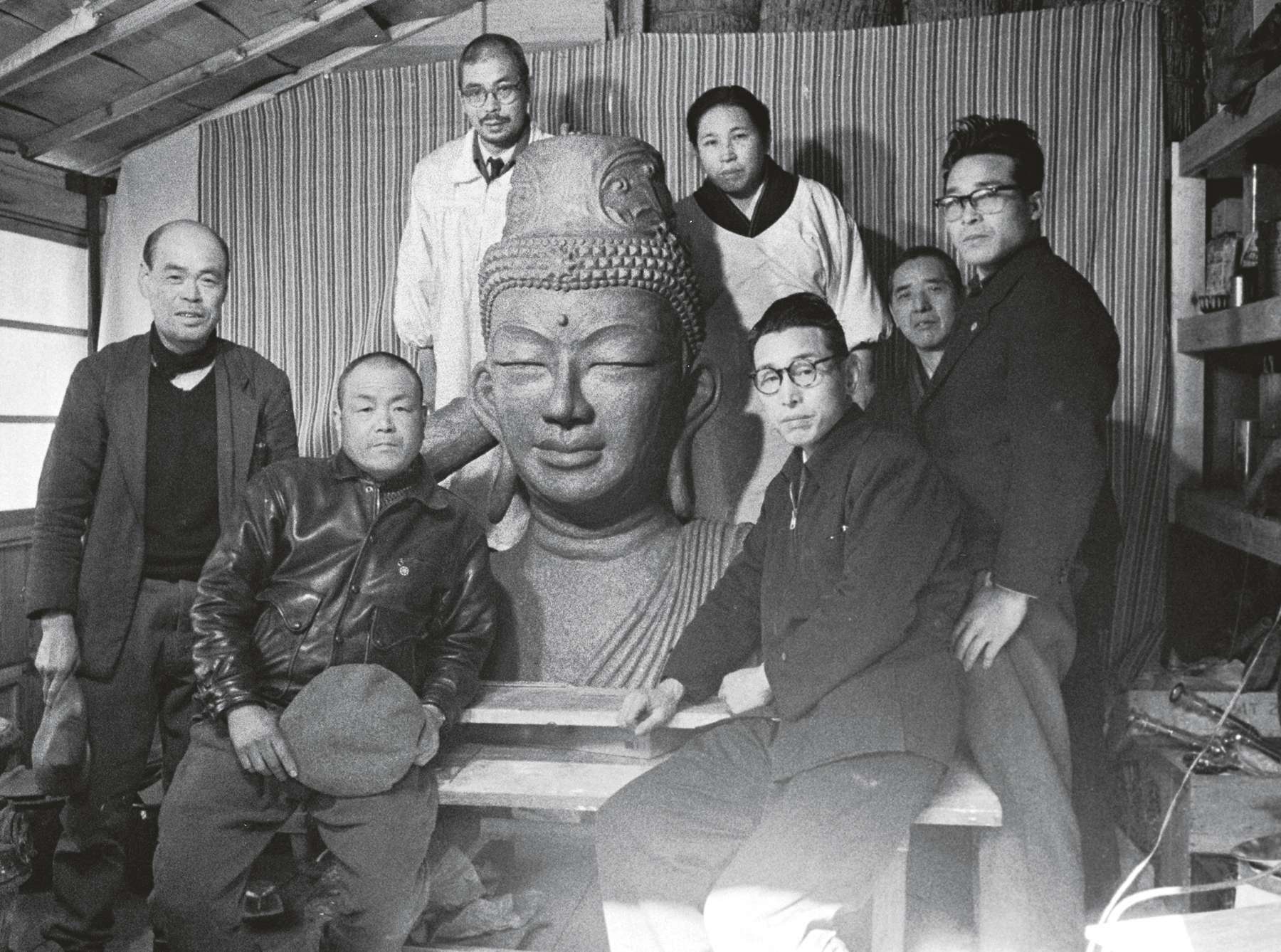 Shinjo and Tomoji, both wearing smocks over their clothing, stand in a studio behind a large sculpted bust of a smiling buddha, with a group of five men, dressed in lay clothes, arranged in front.