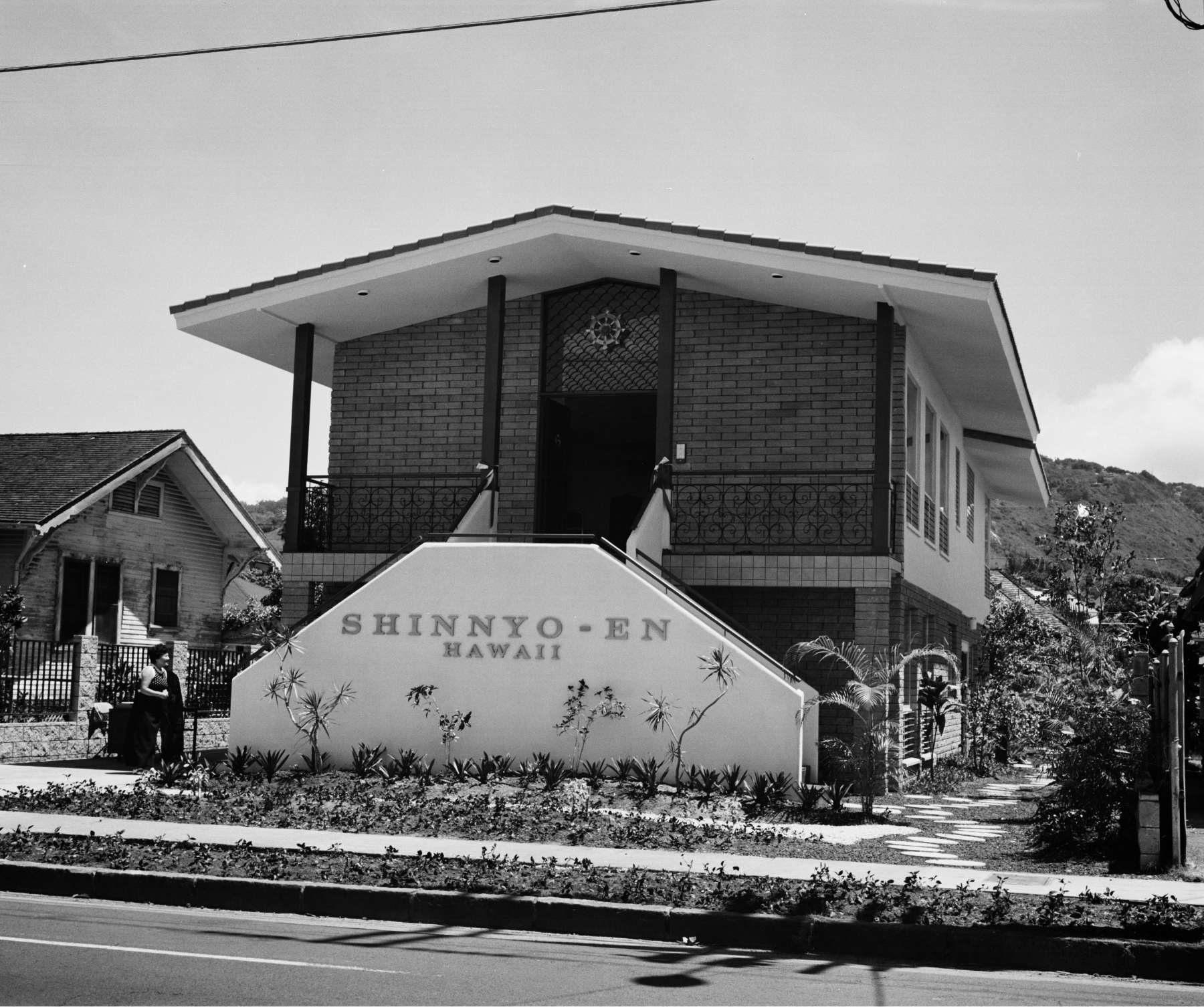 A large modern building with a peaked roof overhanging a two story portico approached by divided staircases, the front of which bears the words “Shinnyo-en Hawaii.”