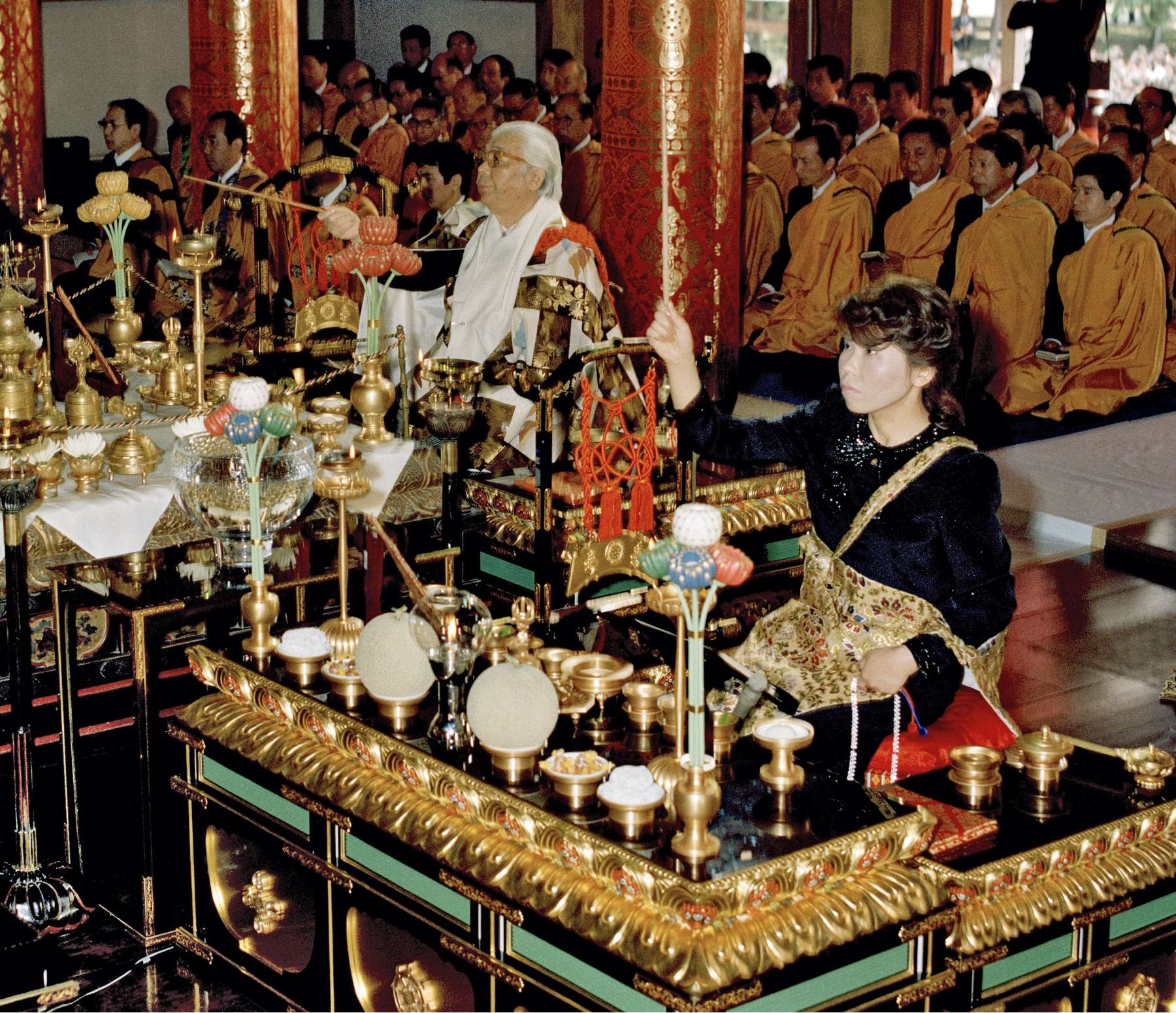 Shinjo, white haired, in brocade robes, and Masako, in formal dress, both seated before ornate tables filled with ritual materials, jointly conduct a rite in a temple filled with monks in formal robes.