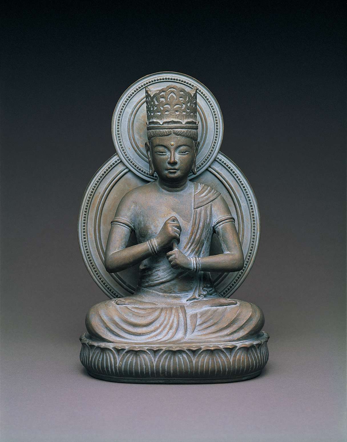 A matte, blue and gray hued  statue of a buddha wearing a tall crown, sitting cross-legged atop a seat of curving lotus petals, right hand grasping upturned left index finger in front of the body.