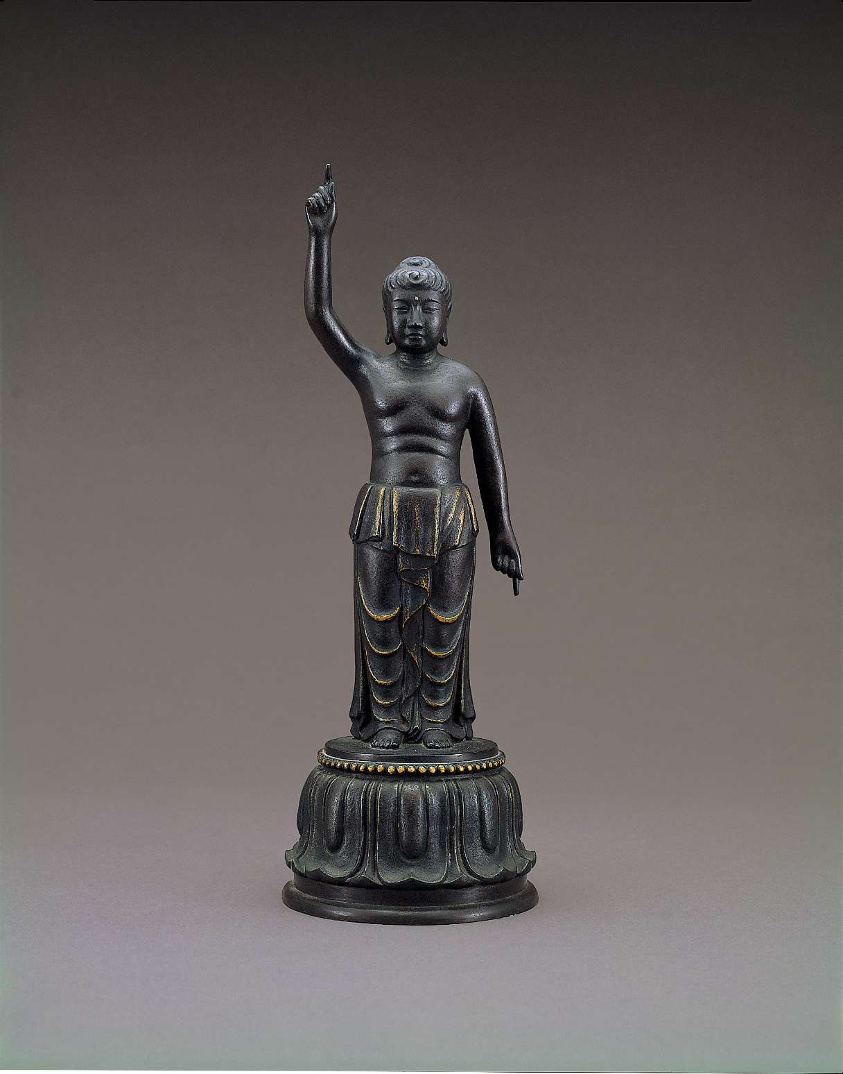 A small, dark-colored matte statue of Buddha, upper body unclothed, standing atop a lotus pedestal, right hand pointing at the sky, left at the ground; his lower garment drapes down the legs in waves.