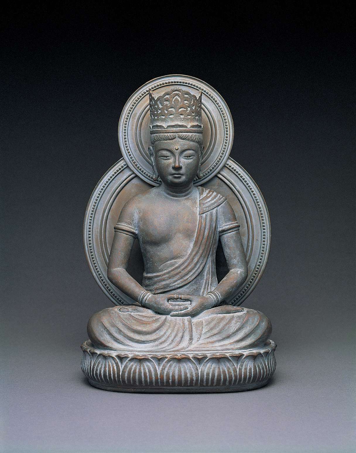 A matte, blue and gray hued  statue of a buddha wearing a tall crown, sitting cross-legged atop a seat of curving lotus petals, hands in meditation, with a circular nimbus behind his body and head.