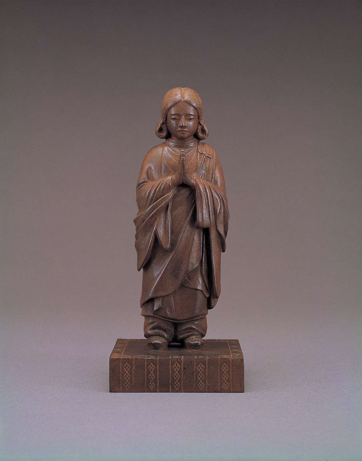 A matte brown statue of a long-haired young child, wearing flowing, oversized robes that accentuate his diminutive stature, stands with palms pressed together at his heart atop a decorated pedestal.