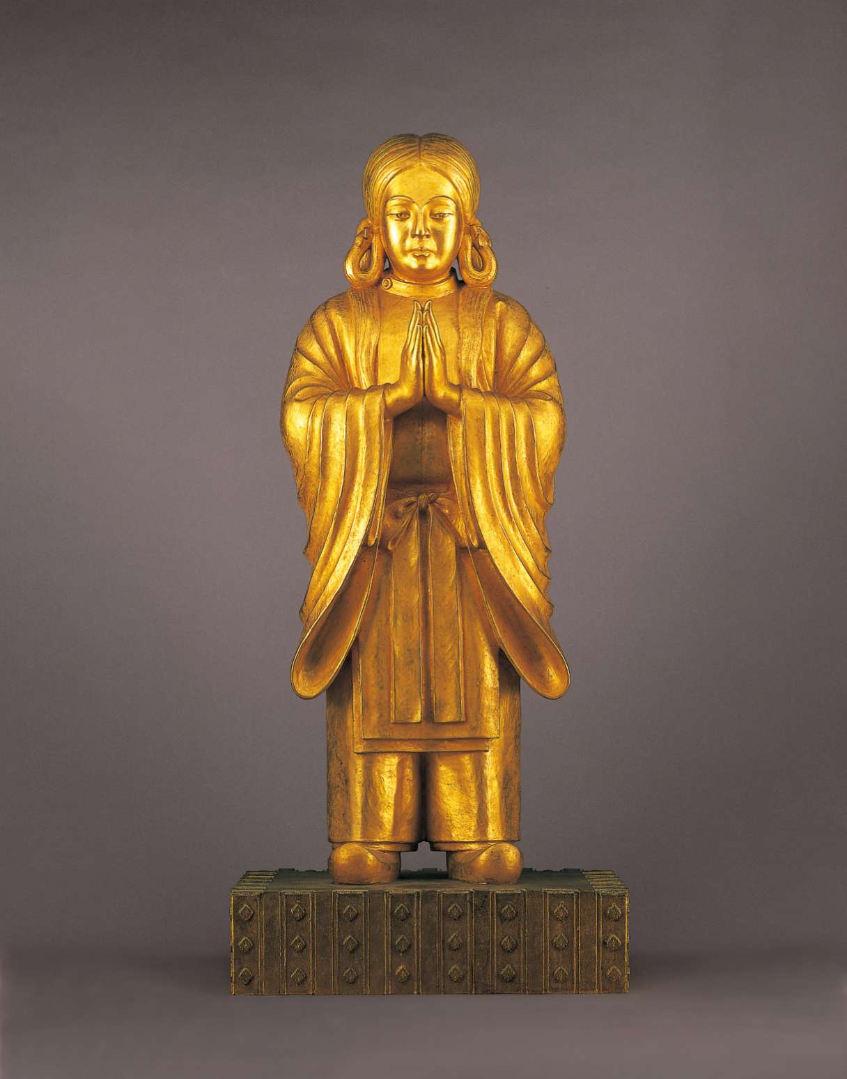 A lustrous, bronze hued statue of a long-haired youth, wearing a kimono with flowing sleeves, an apron, stiff pants, and shoes with upturned toes stands with hands in prayer atop a decorated pedestal.
