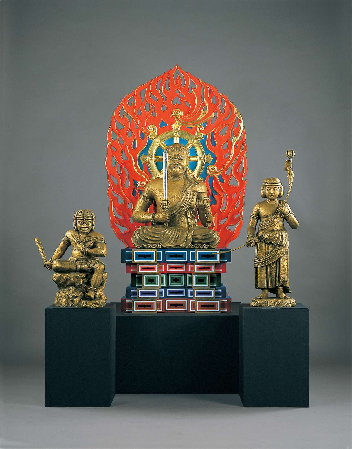 A statue of a scowling figure sitting atop a tall, variegated throne, holding an upturned sword in his right hand and noose in his left, backed by a golden wheel and a brilliant red halo of flame.