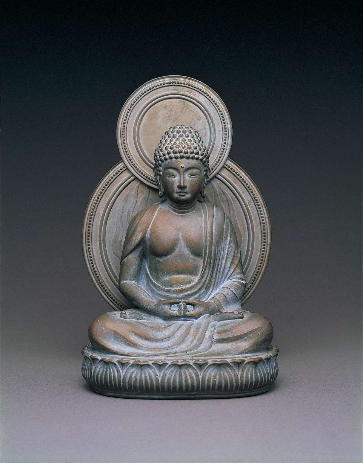 A matte, gray hued statue of a buddha sitting cross-legged atop a seat of curving lotus petals, with a circular nimbus behind his body, hands in meditation.