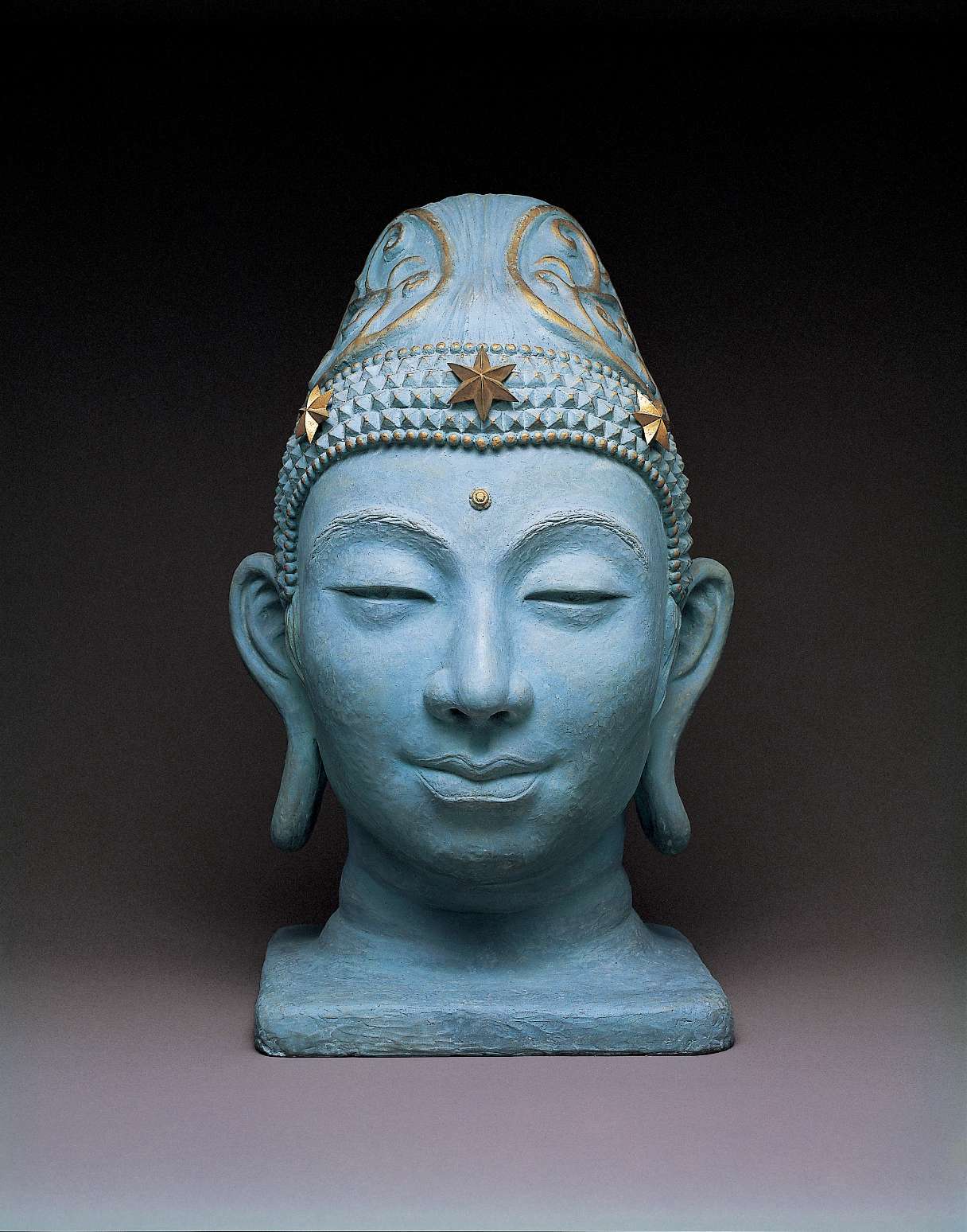 A light-bluish hued bust of a slightly smiling Buddha wearing a decorative headpiece embellished with golden highlights; the face looks like a person one might see, rather than a stylized icon.