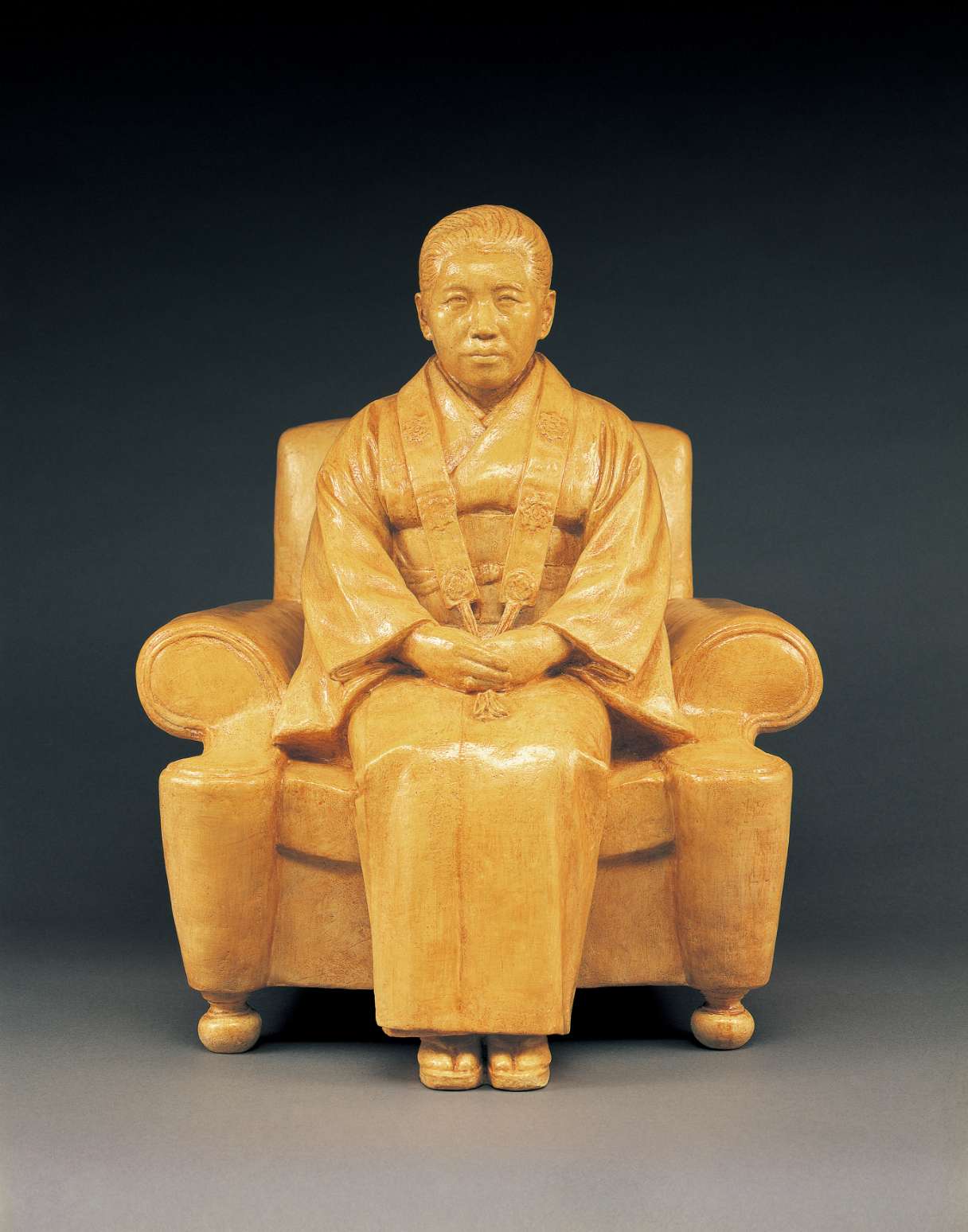 An ochre hued statue of a middle-aged Japanese woman, hair pulled back, wearing a kimono draped with kesa, seated in an upholstered chair, her hands folded neatly in her lap, feet flat on the ground.