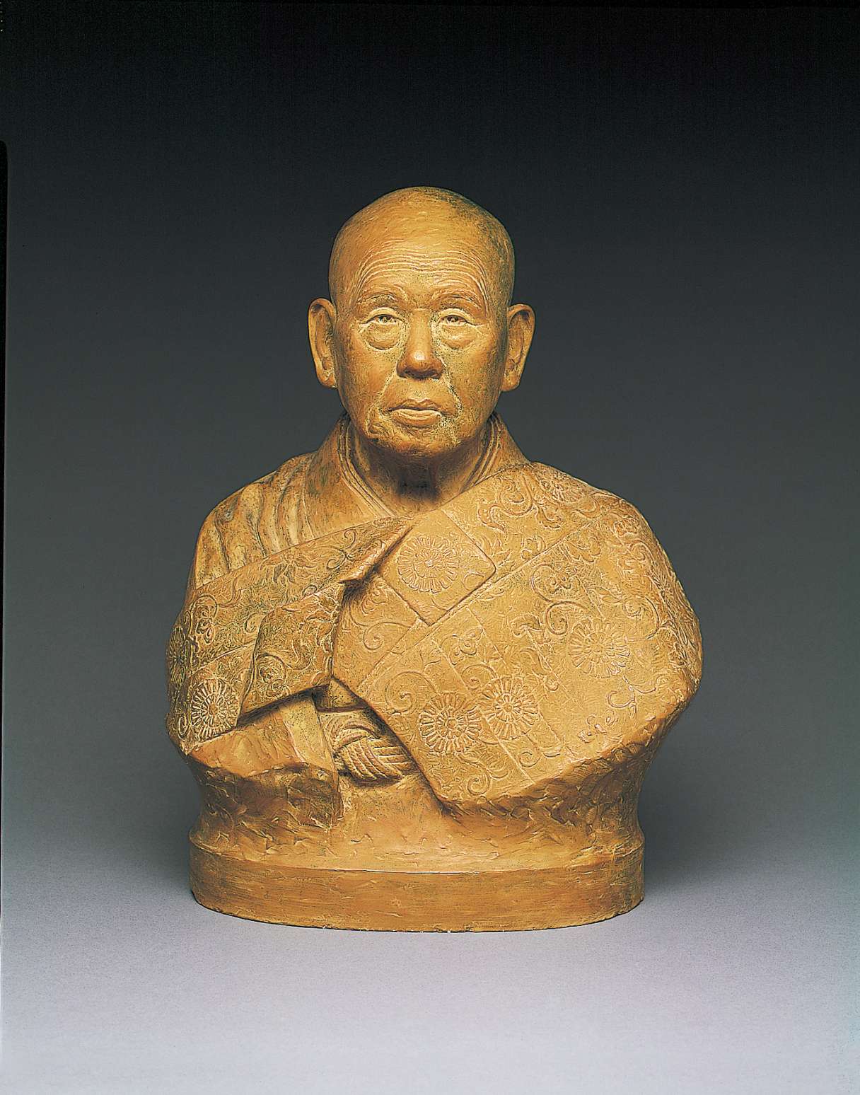 An ochre hued bust of a shaven headed, elderly Japanese man, with kind eyes and large ears, his face wrinkled with age, wearing a kimono over which is draped an ornately decorated patched robe.