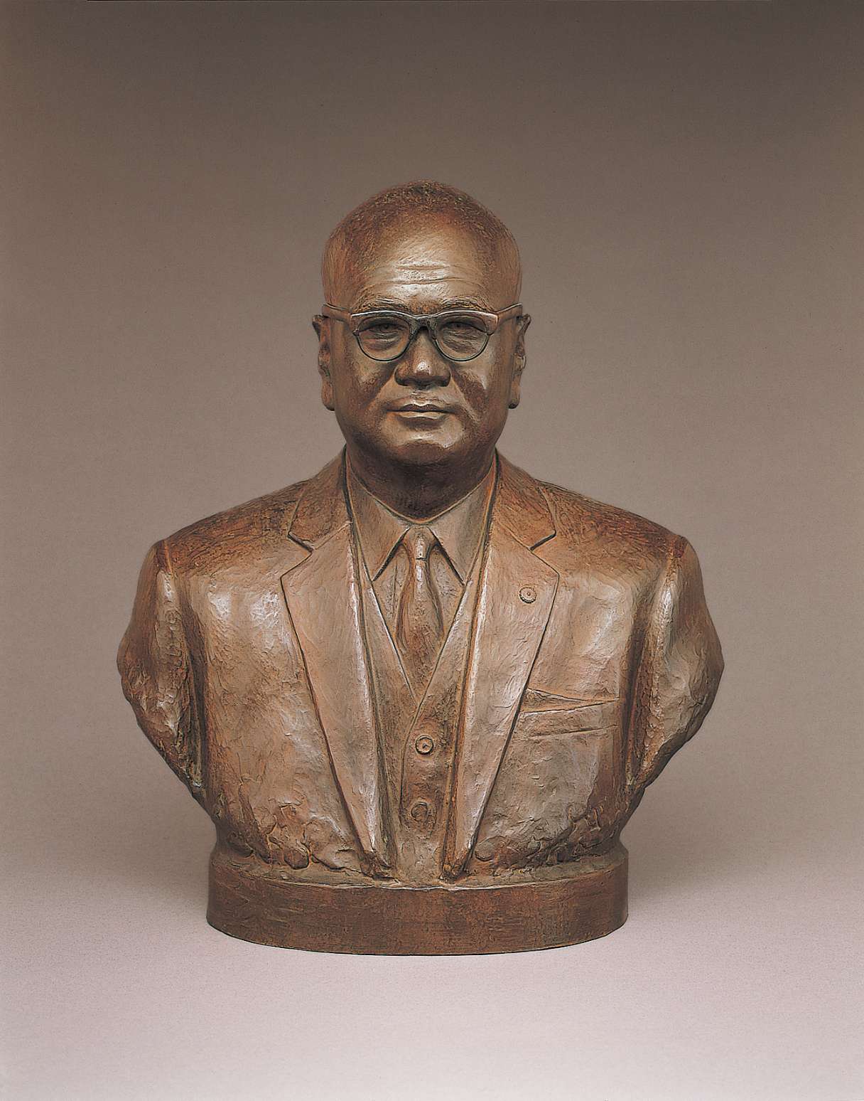 A brown hued bust of a bald, spectacled, middle-aged Japanese man wearing a suit and tie, his full cheeks, eyes, and mouth smiling slightly, with a few wrinkles visible on his forehead.
