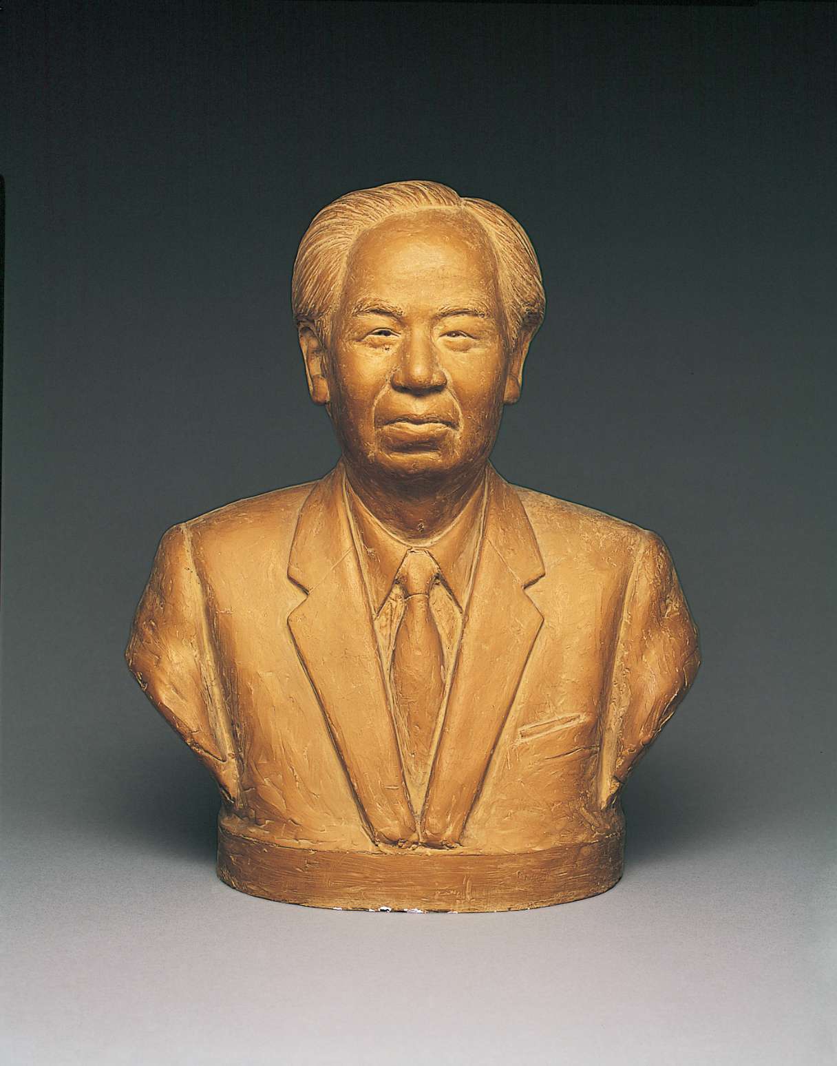 An ochre hued bust of a Japanese man wearing a suit and tie, with medium length hair parted to the left above a receding hairline and a friendly face with smiling eyes and a slight smile on the lips.