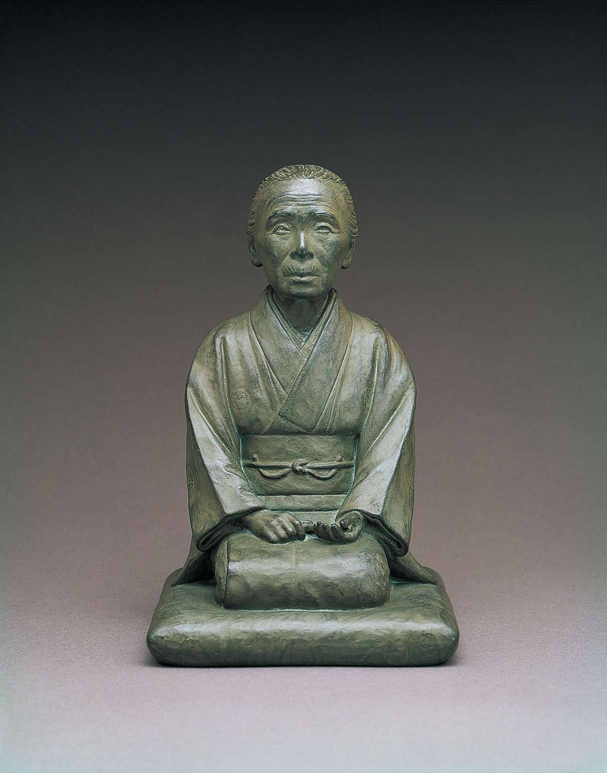 A gray, metallic statue of an elderly Japanese woman, hair pulled back, wearing traditional kimono and kneeling on a cushion. Her hands rest in her lap and her attentive face seems as if listening.