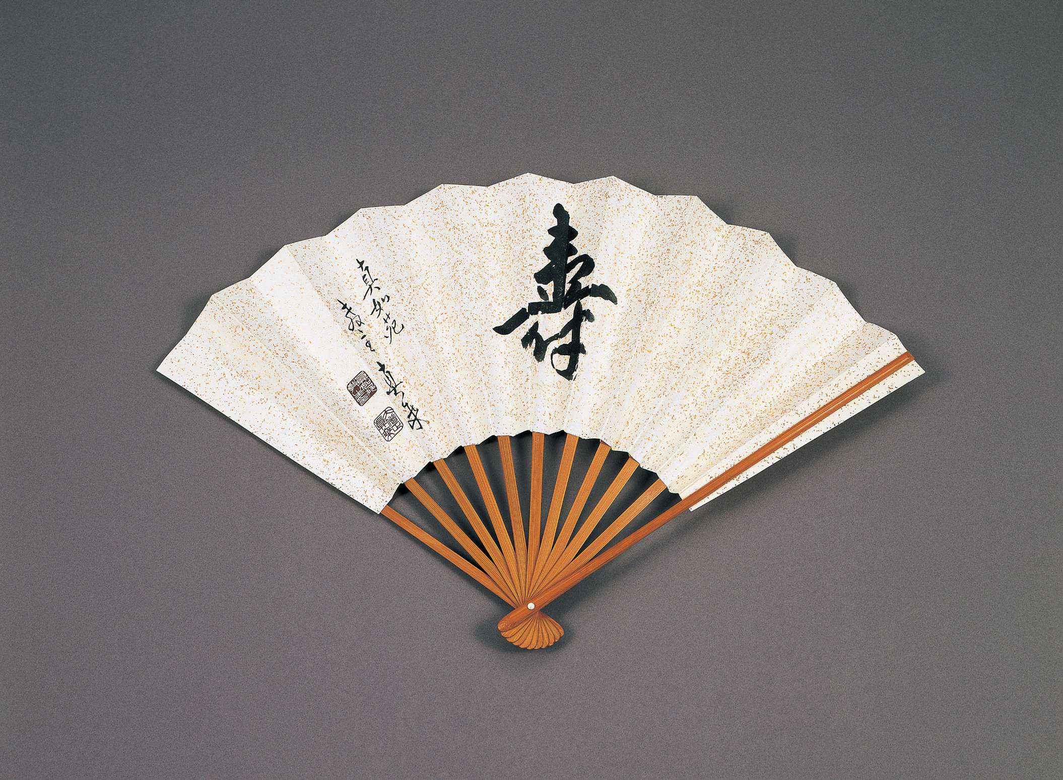 A large Japanese character is written with brush and ink on an opened paper folding fan. Two smaller lines of calligraphy are written to the left, with two black stamp impressions.