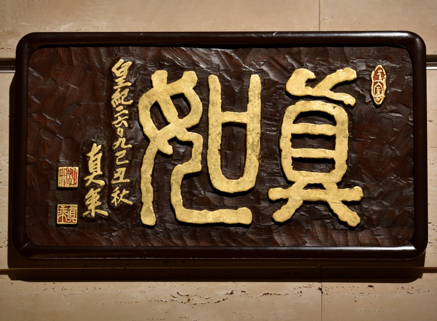 A dark brown slab of wood is carved with stylized, thick lined Japanese characters in gold, embellished with nearby vertically aligned calligraphy in gold and red and gold calligraphic stamps.
