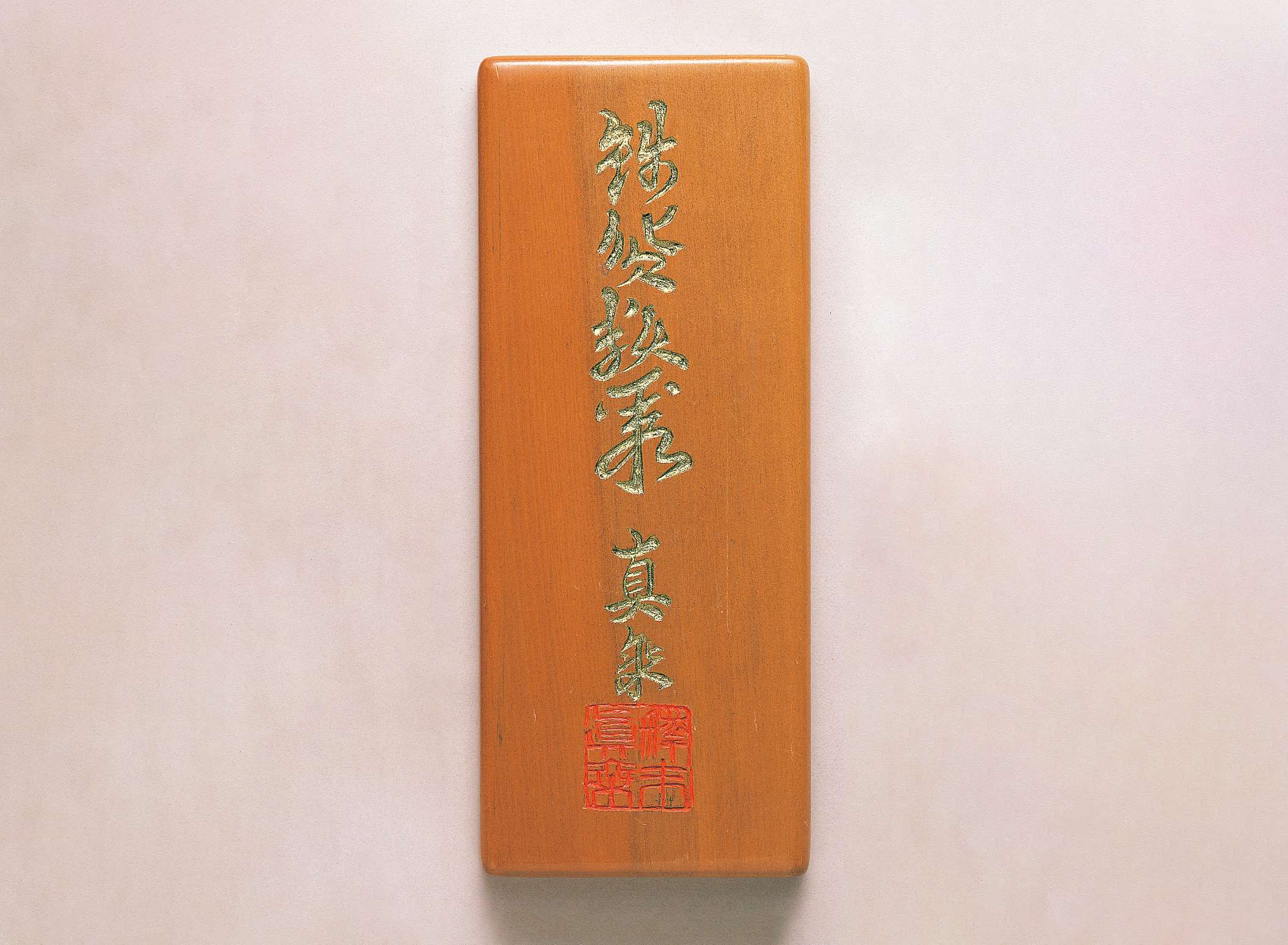 A vertically oriented rectangular slab of smooth, polished sienna colored wood is etched with flowing Japanese calligraphy in gold, beneath which a deep red calligraphic seal is etched.