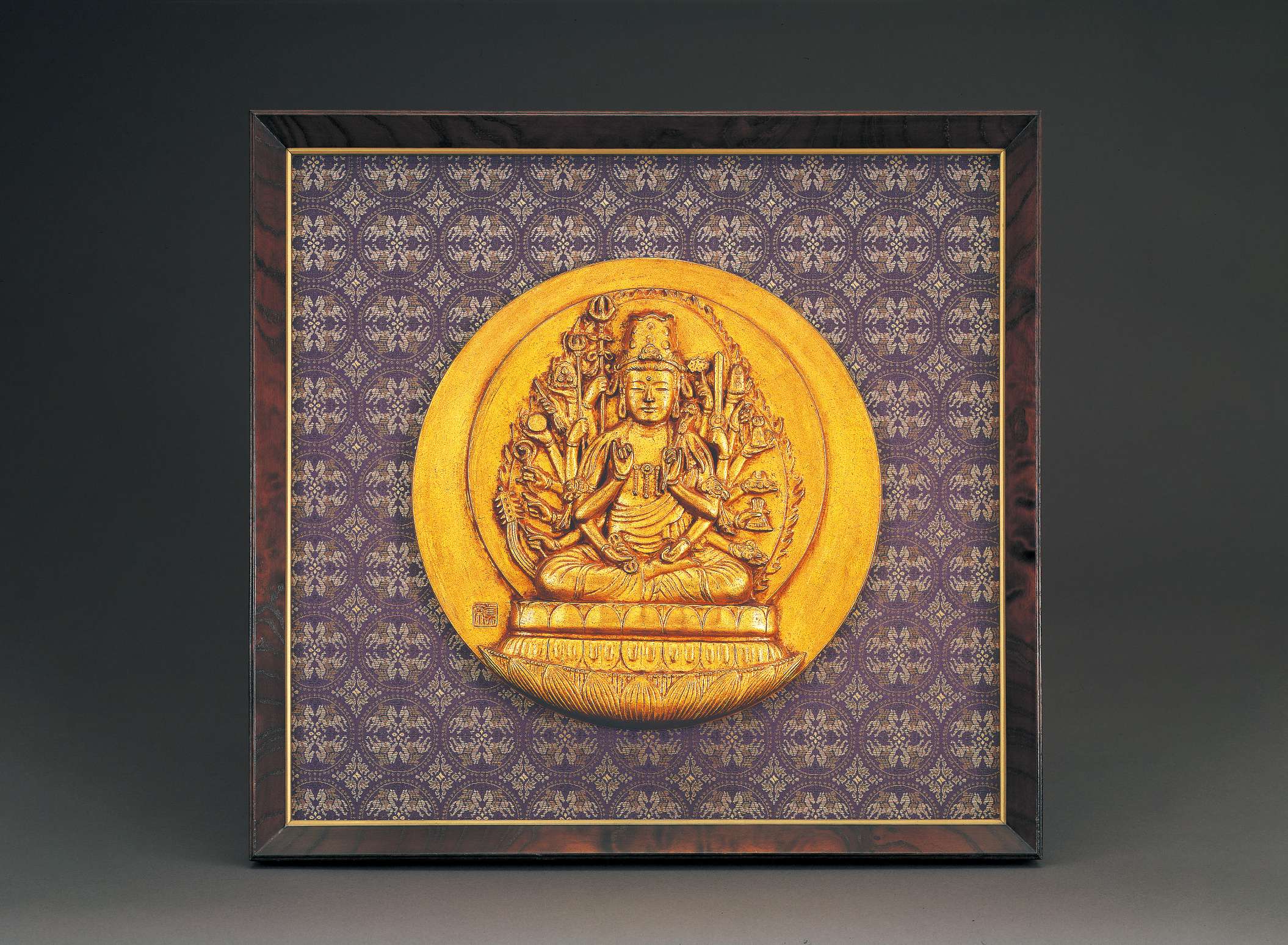 A circular golden relief of a bodhisattva in royal garb and jewelry, wearing a high crown, with an array of twenty arms bearing symbolic implements, sitting cross-legged atop a stylized lotus seat.