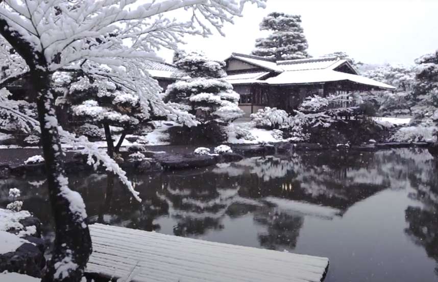 Snow covered trees surround a Japanese temple set beside a reflecting pond.