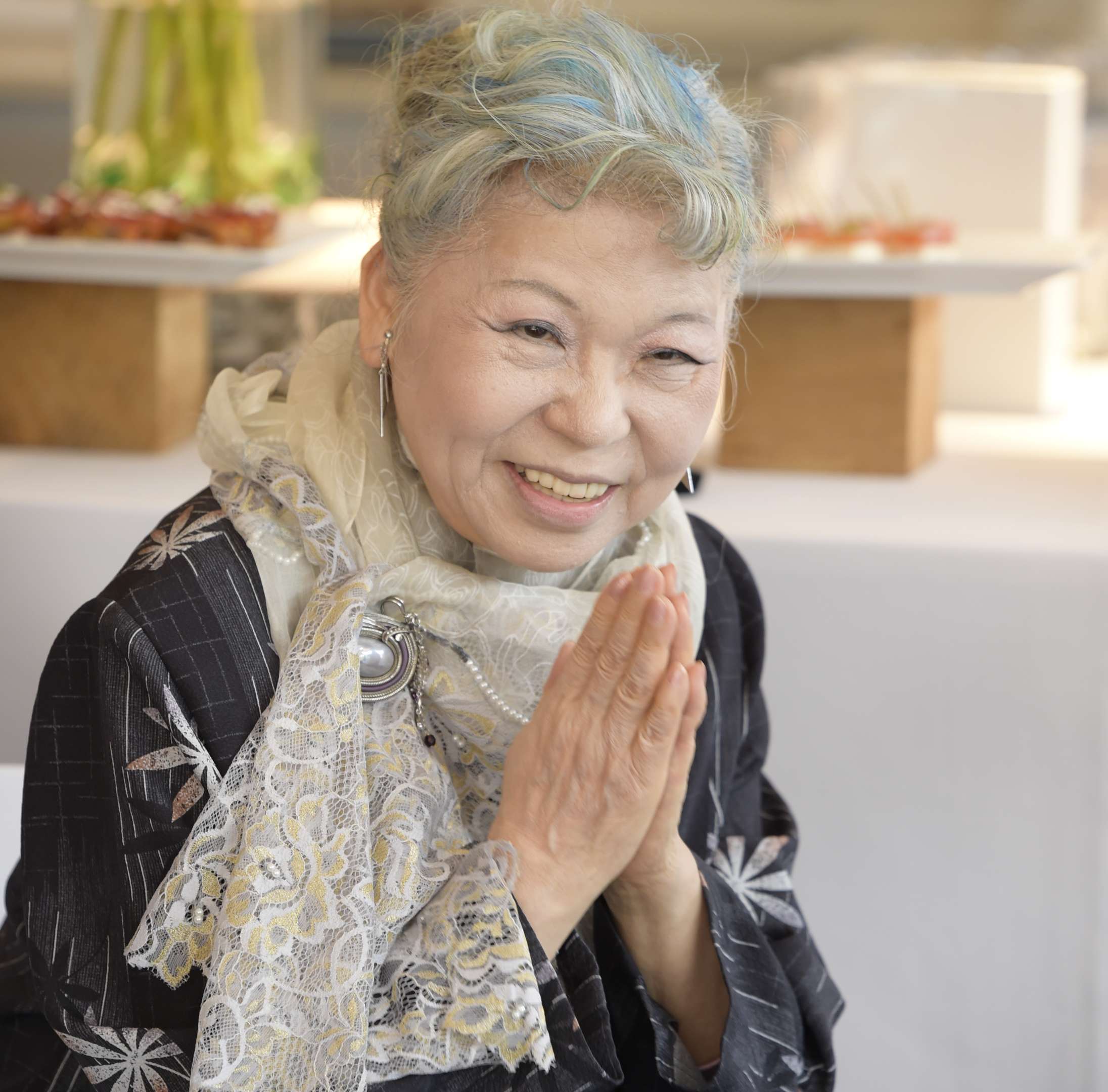 A photograph of Her Holiness, gray-haired, wearing a lace scarf, and offering a warm smile with hands in gassho.