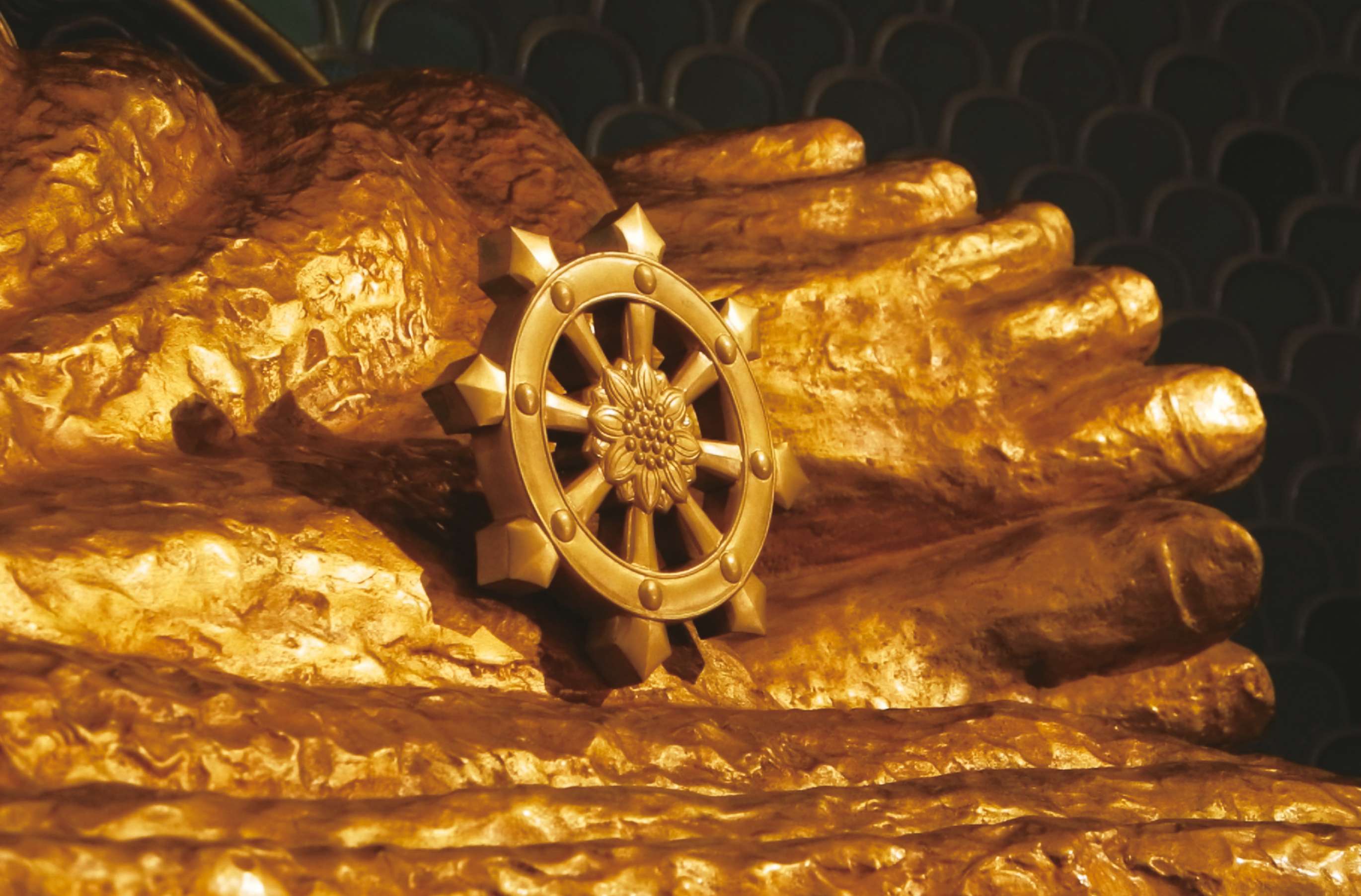 A photograph at close range of the feet of a large, golden Nirvana Buddha statue, showing in detail the hand-sculpted surface of the feet and a Dharma wheel placed in front of them.