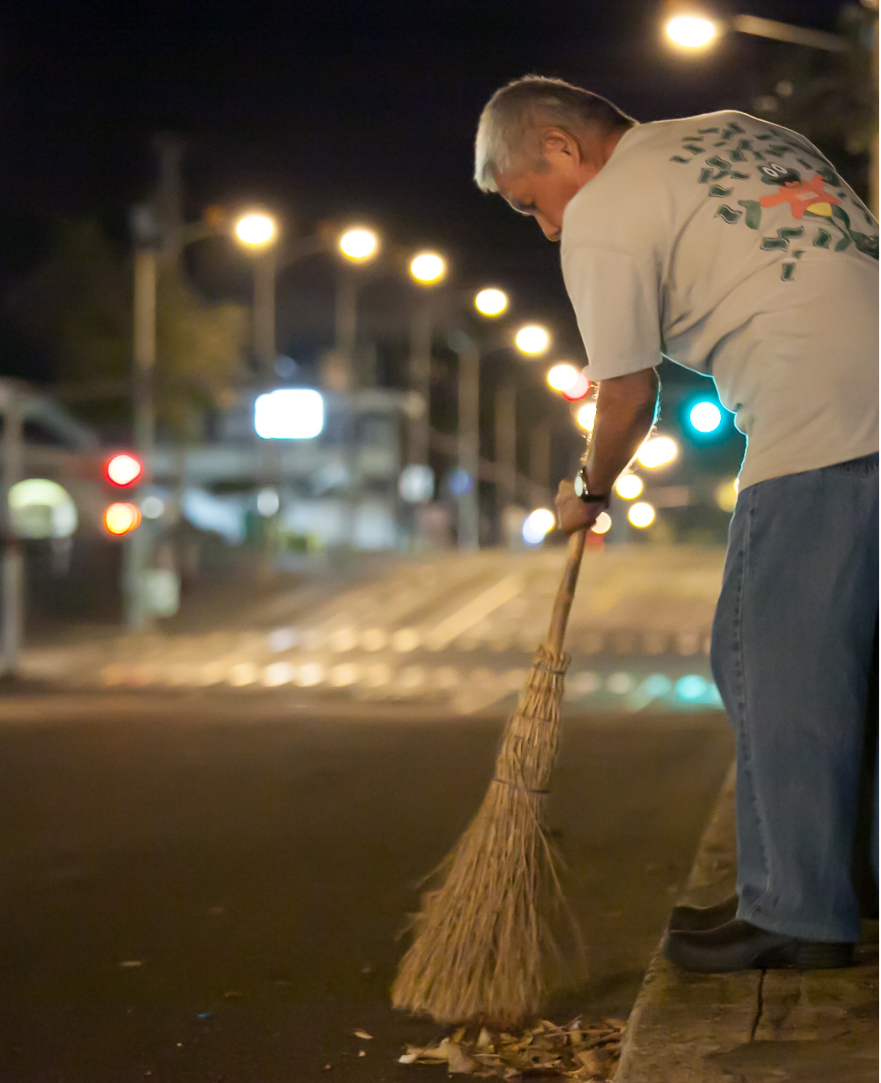 On a city street in the predawn darkness, a gray-haired man sweeps trash into a neat pile near a curb.