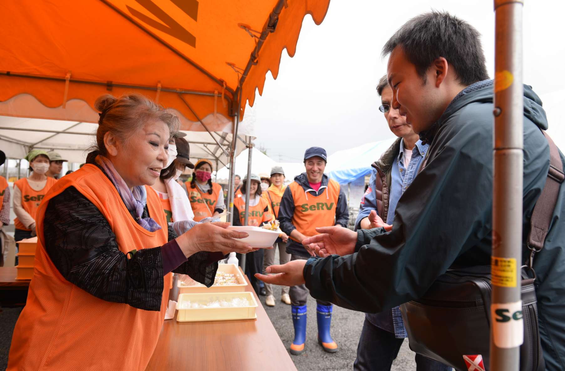 Her Holiness, standing beneath a temporary awning, wearing an orange volunteer vest over her jacket, offers a bowl of soup to a man who reaches out to accept it.