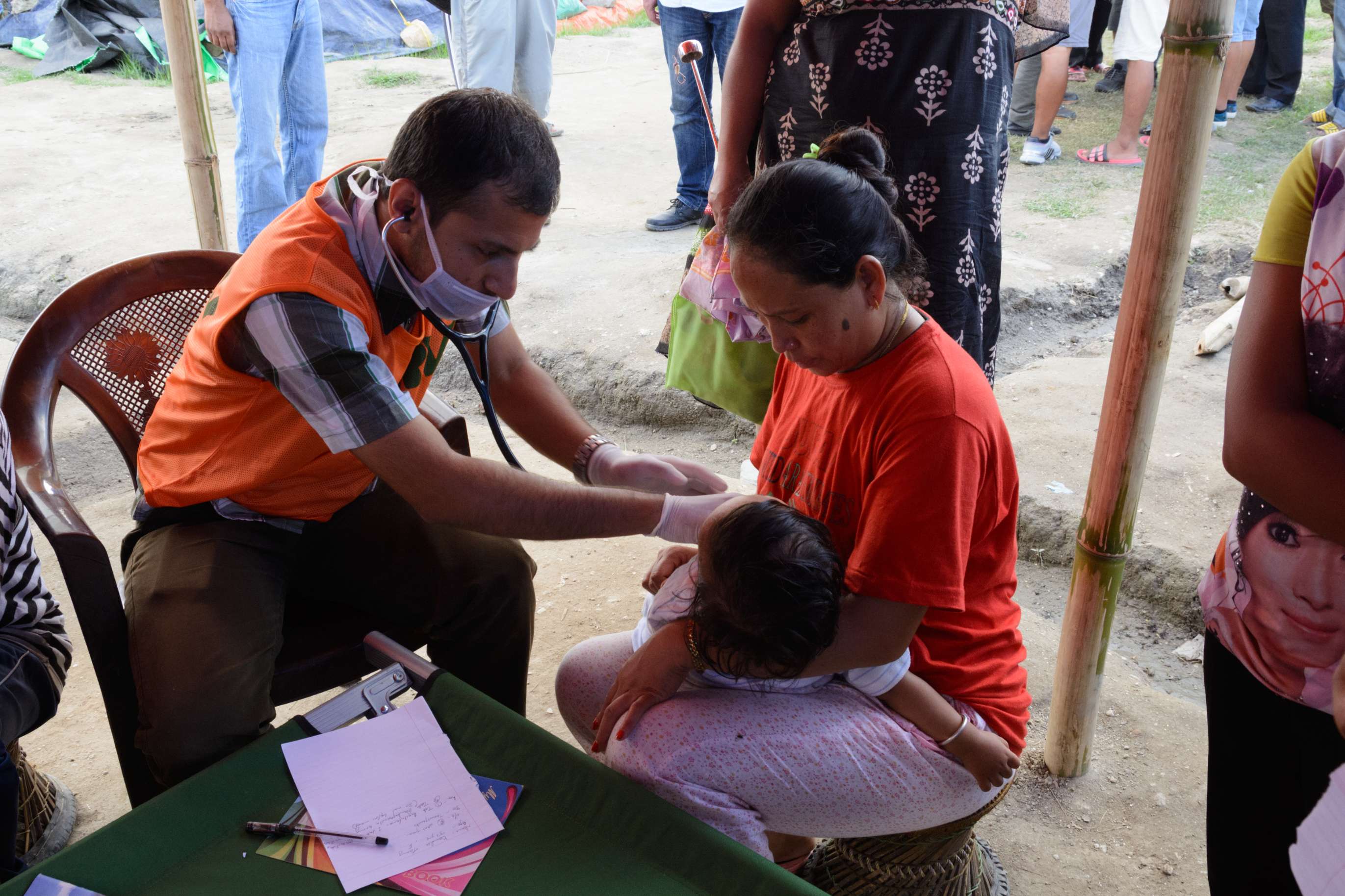 A doctor in an orange vest uses a stethoscope to examine a child held in the arms of a woman at an outdoor clinic.