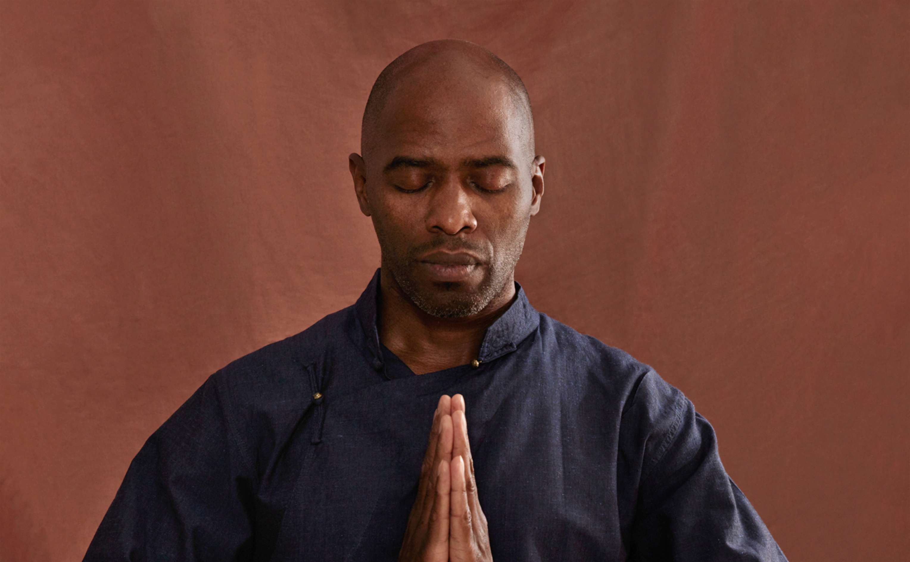 A man with eyes closed in a contemplative state, presses his palms together in front of his chest in a prayerful salutation.