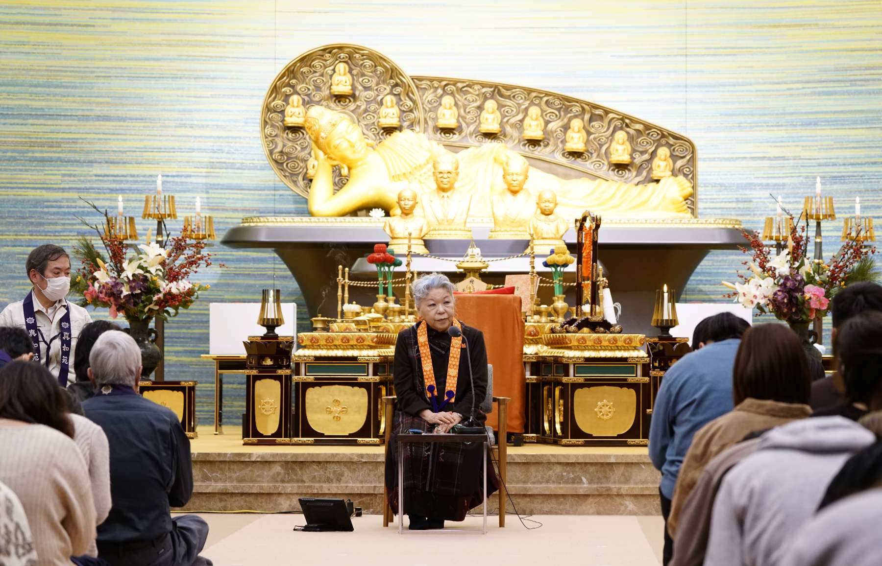 An elderly Japanese woman dressed in black attire with a priestly Buddhist surplice around her shoulders sits on a low chair in front of a Buddhist shrine with a large statue of a reclining Buddha, speaking intently to an audience of Japanese people seated neatly in rows on the floor.