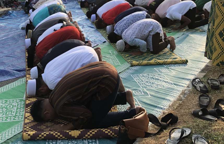 Rows of men wearing long shirts and skull caps bowing on prayer carpets with their heads, hands, and feet touching the ground; discarded sandals can be seen nearby.