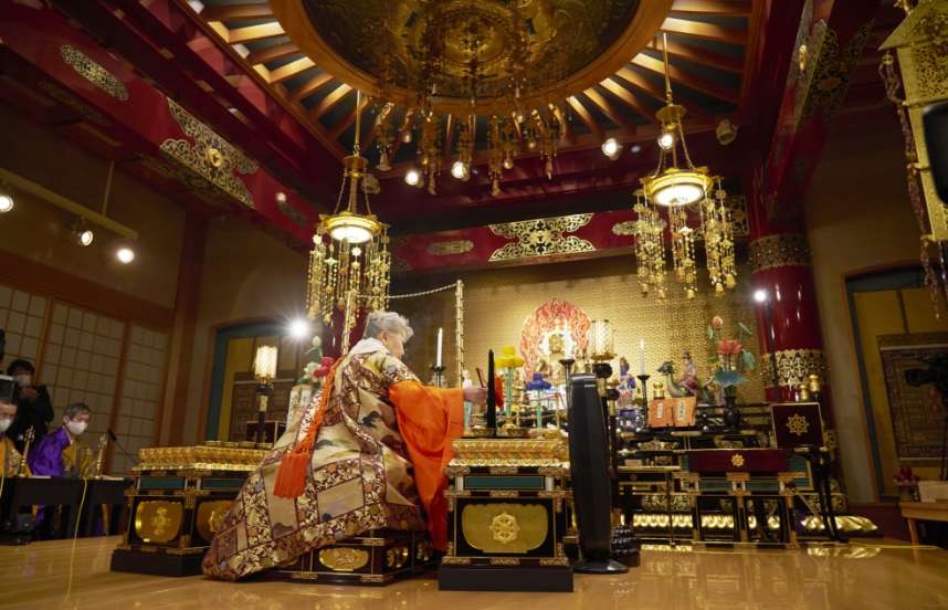 Her Holiness sits before a fire ceremony altar with a large flame and a statue of Fudo-myo holding an implement in her right hand, performing a rite.