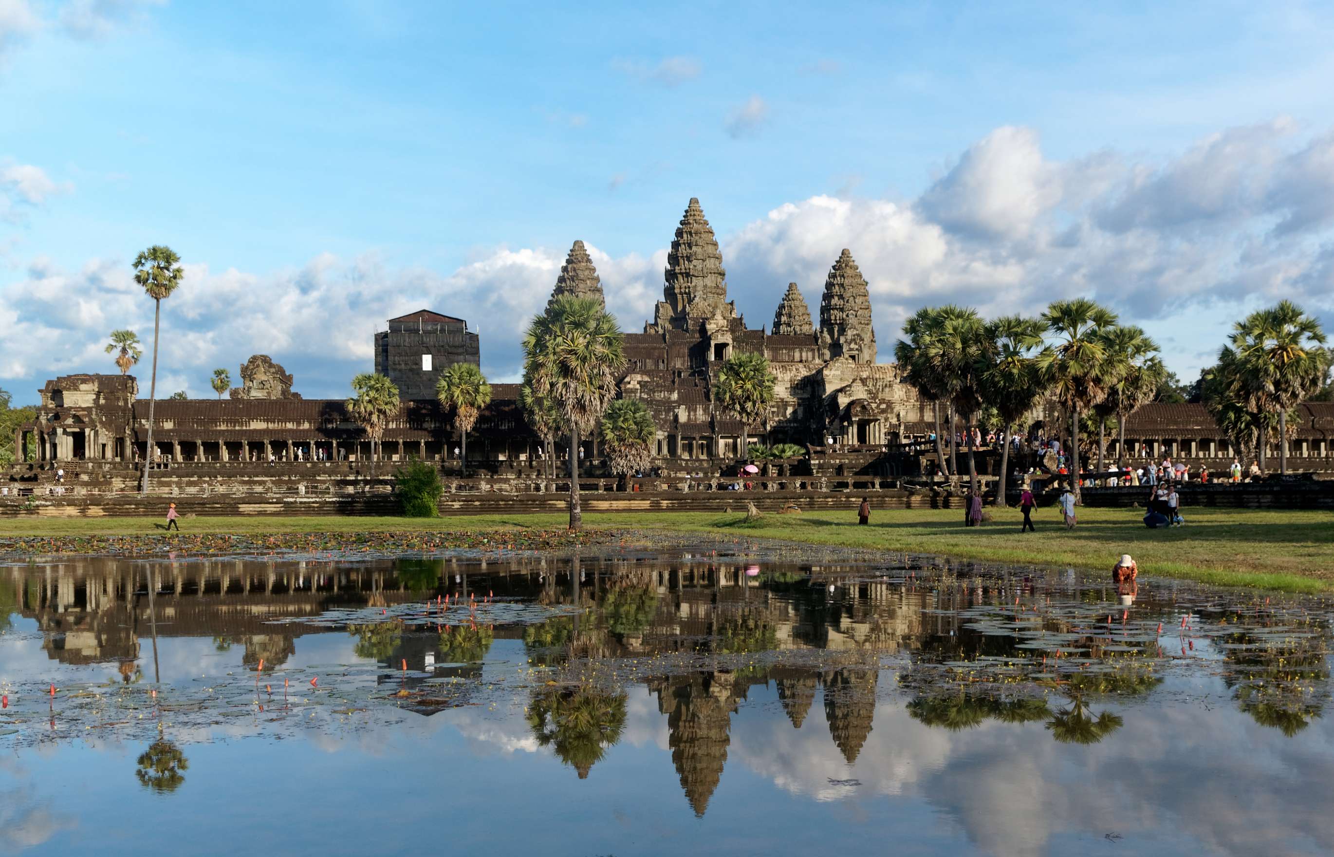 A large, ancient Cambodian temple ruin with long, single storey stone arcades arranged around high conical stone towers sits under a pale blue sky with a sting of clouds in the distance and palm trees in the foreground. The whole scene is reflected in a serene pool of water.